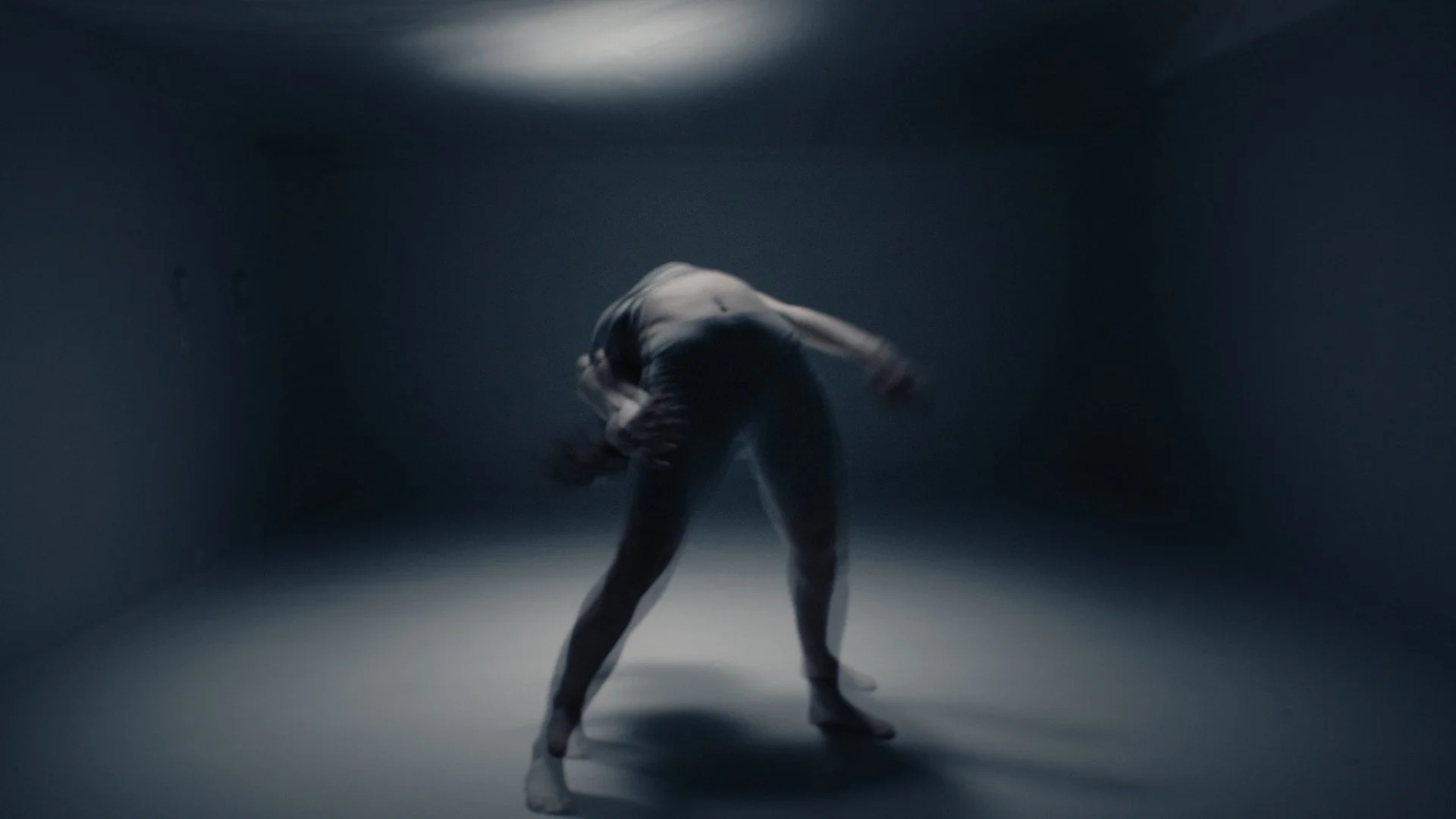 A person performing a dance move in a dimly lit, empty room, with arms bent and body bent forward.