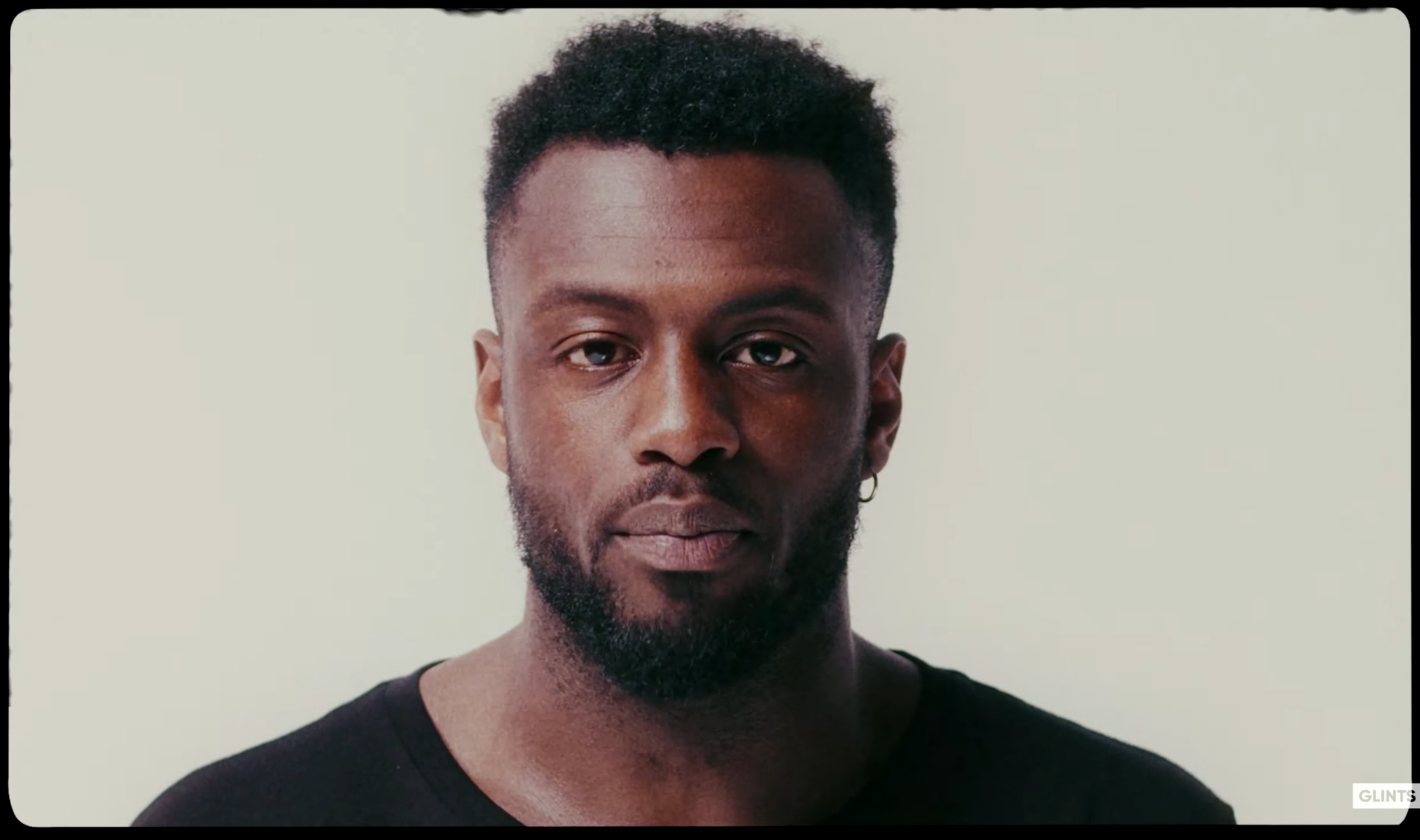 A portrait of a man with dark skin, short curly hair, a beard, a small hoop earring, wearing a black t-shirt, standing against a plain light background.