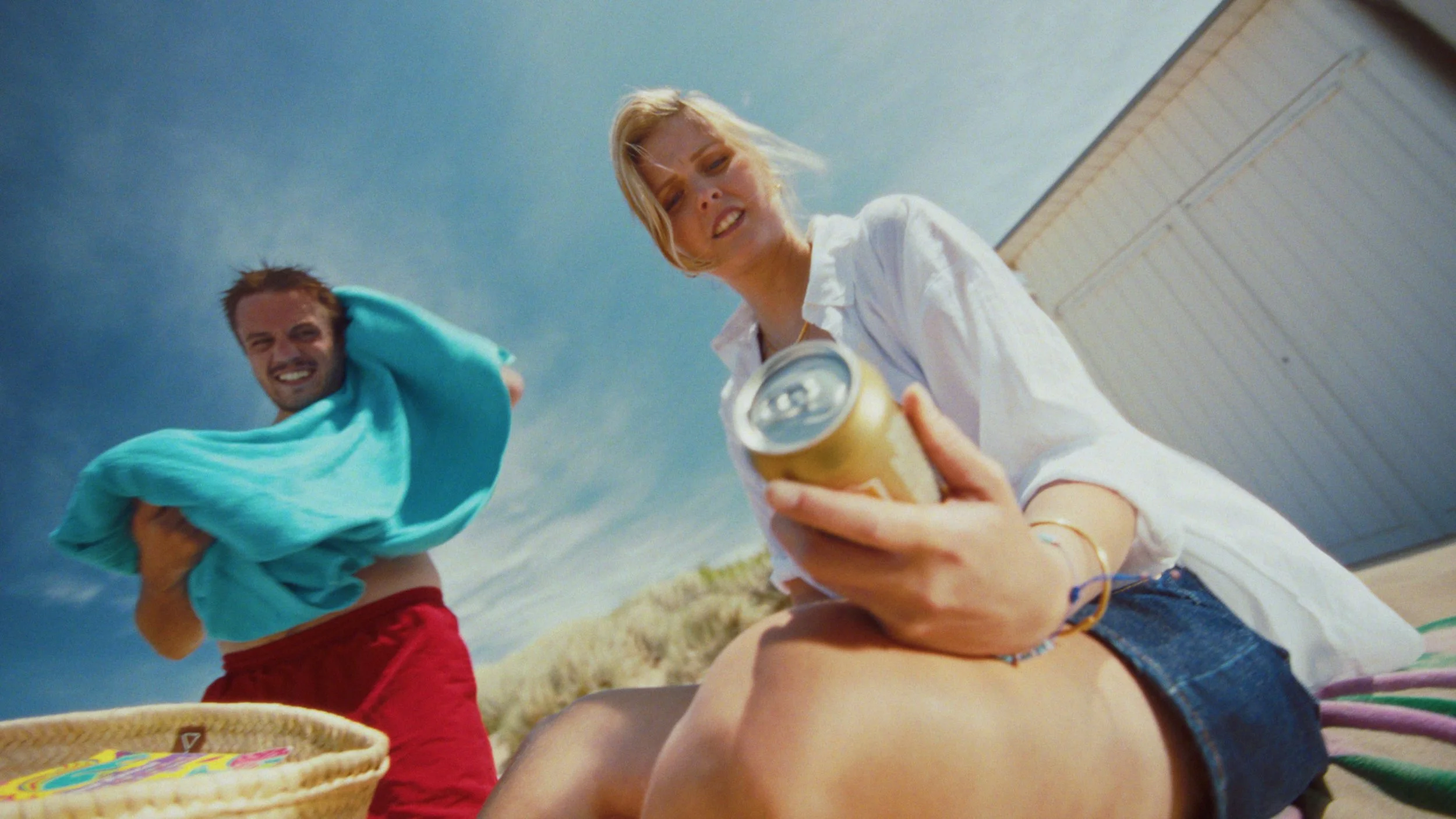 Two young adults enjoying a sunny day outdoors, with the woman sitting on the ground holding a soda can and the man standing nearby with a towel over his shoulder, both smiling.