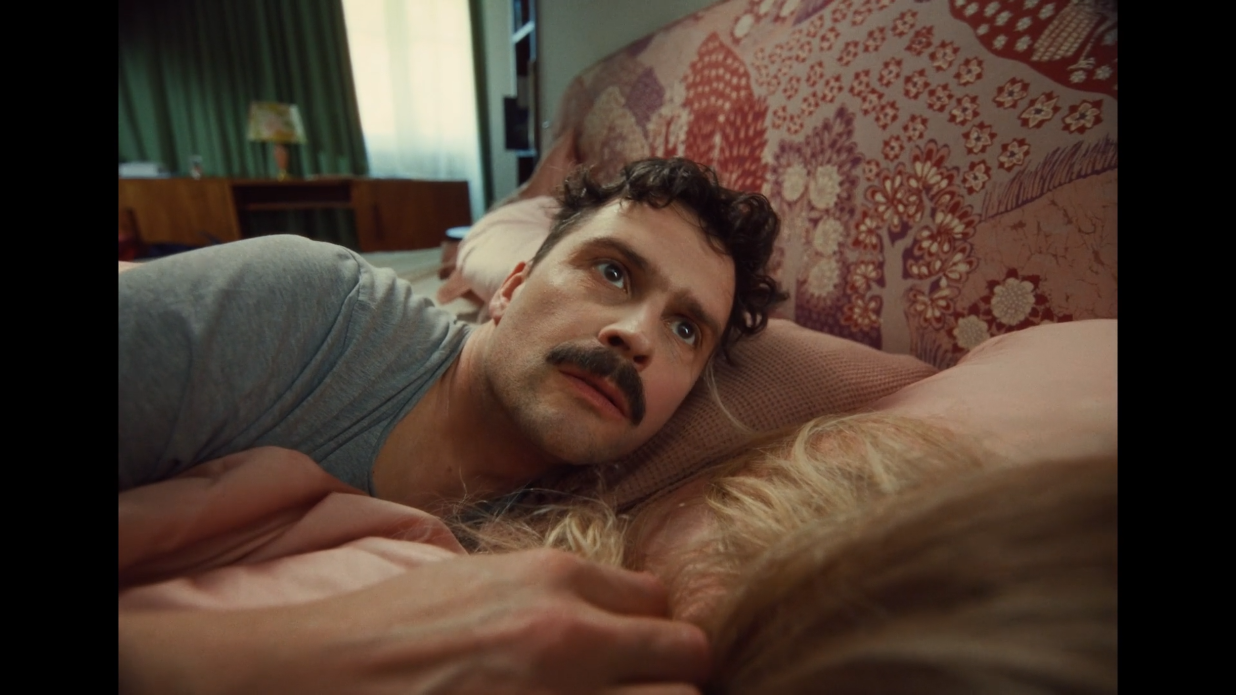 A man with a mustache is lying on his side on a bed, looking up with a serious expression. There is a woman with blonde hair lying beside him, holding his hand. The bedroom has a patterned headboard, curtains, and a wooden nightstand with a lamp.