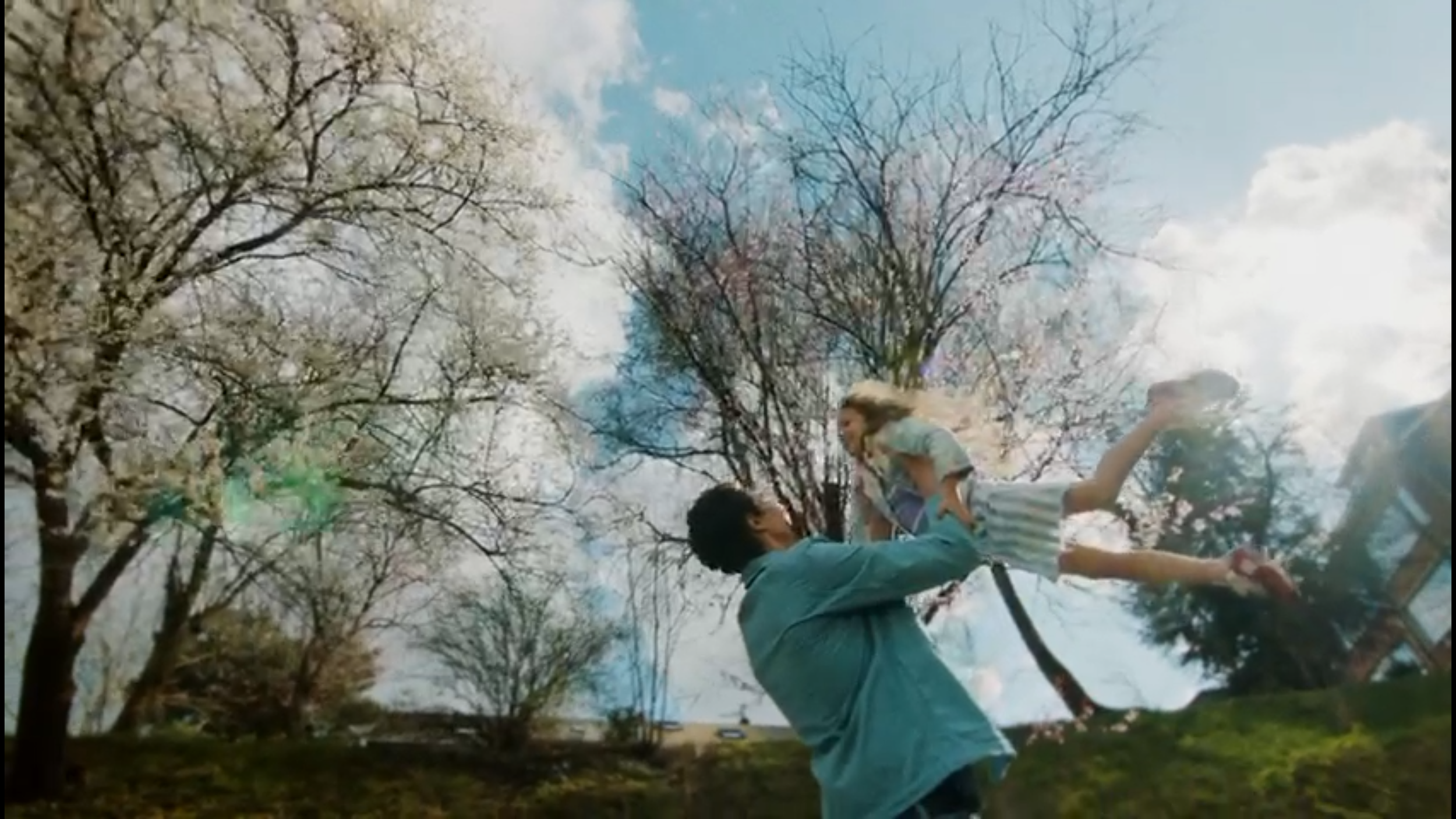 A man lifting a young girl into the air outdoors under a cloudy sky, with blooming trees nearby.