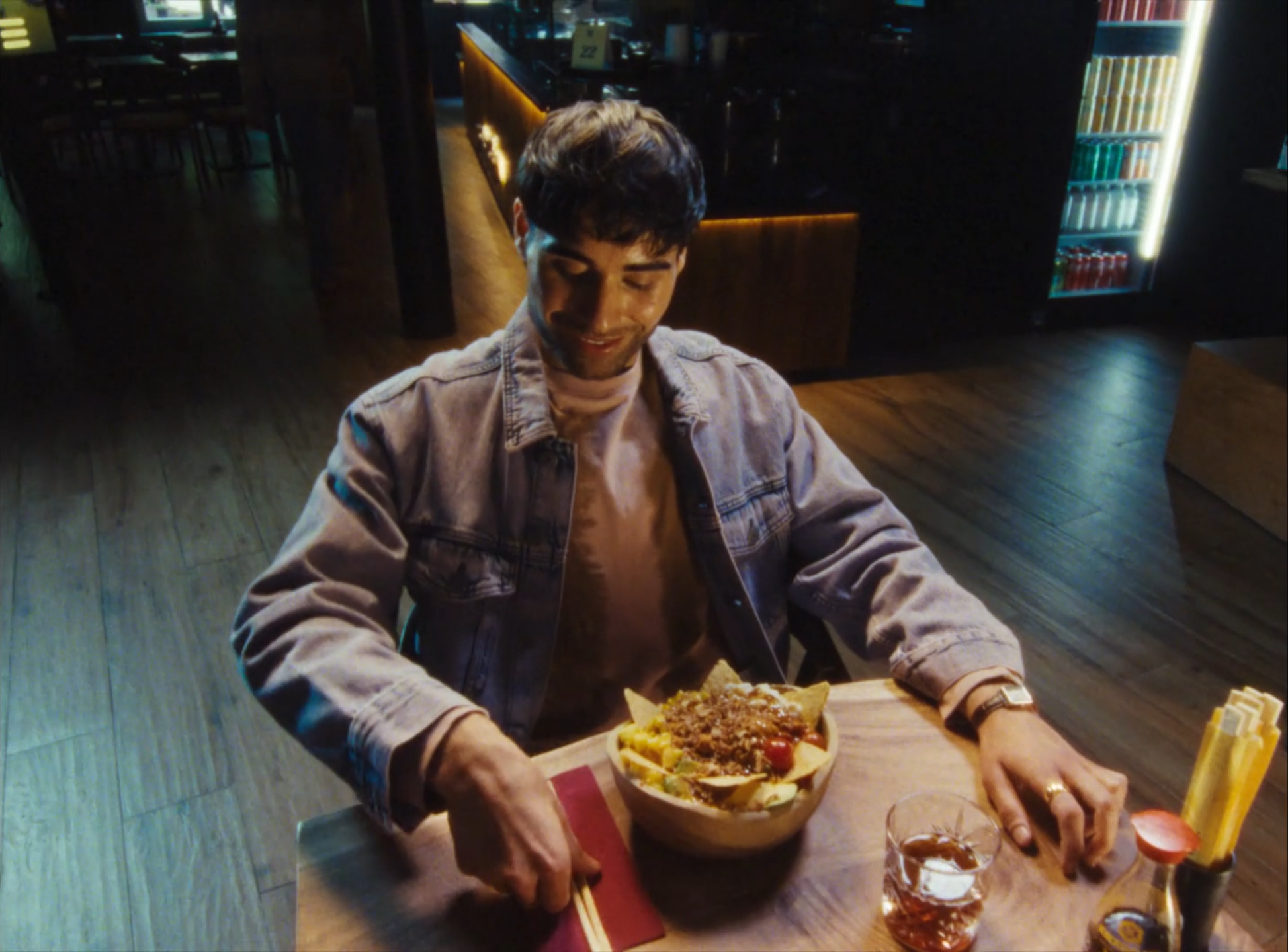 A man sitting at a wooden table in a restaurant, smiling while looking at a bowl of tacos topped with ground meat, with a glass of tea and condiments nearby.