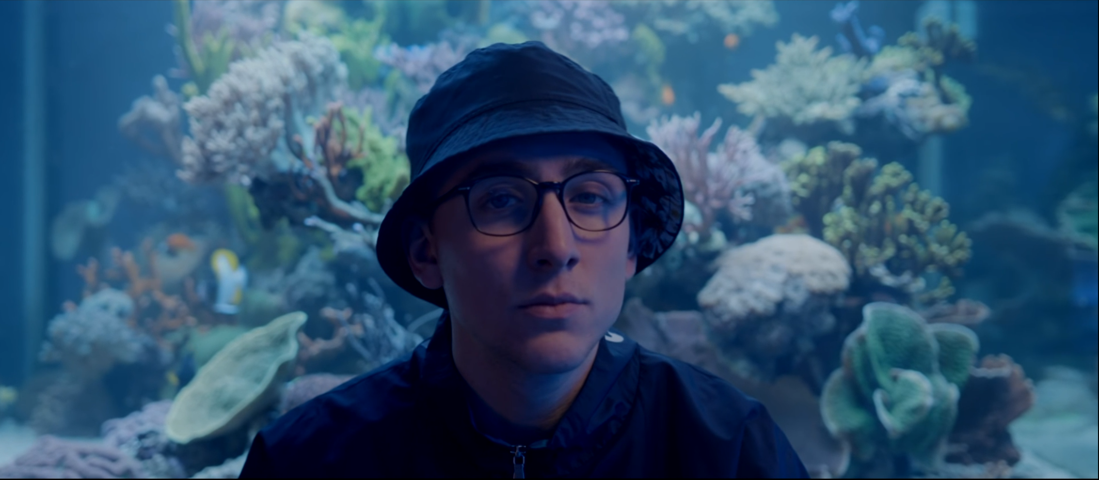 A young man wearing glasses, a bucket hat, and a black jacket stands in front of an aquarium with colorful coral and fish.