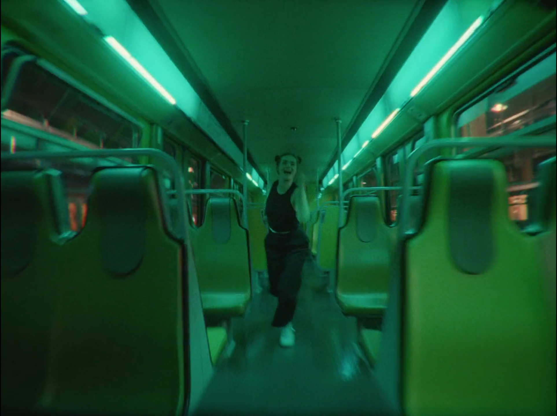 A woman with pigtails jogging or dancing inside a green-lit empty bus at night.