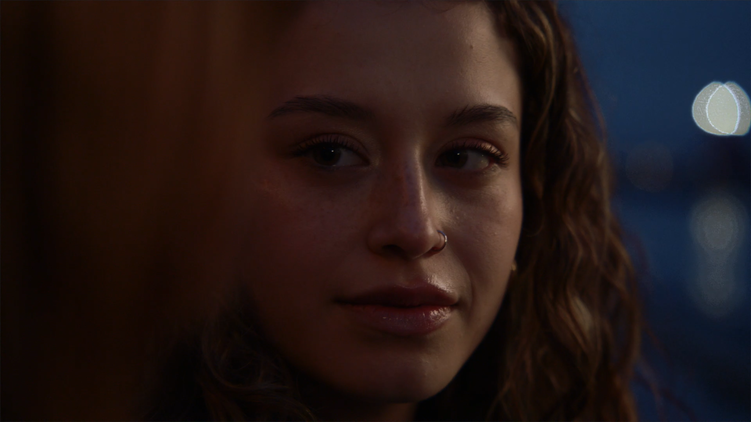 Close-up of a woman's face with light makeup, a small nose ring, and curly brown hair, with a blurred background featuring circular light reflections.