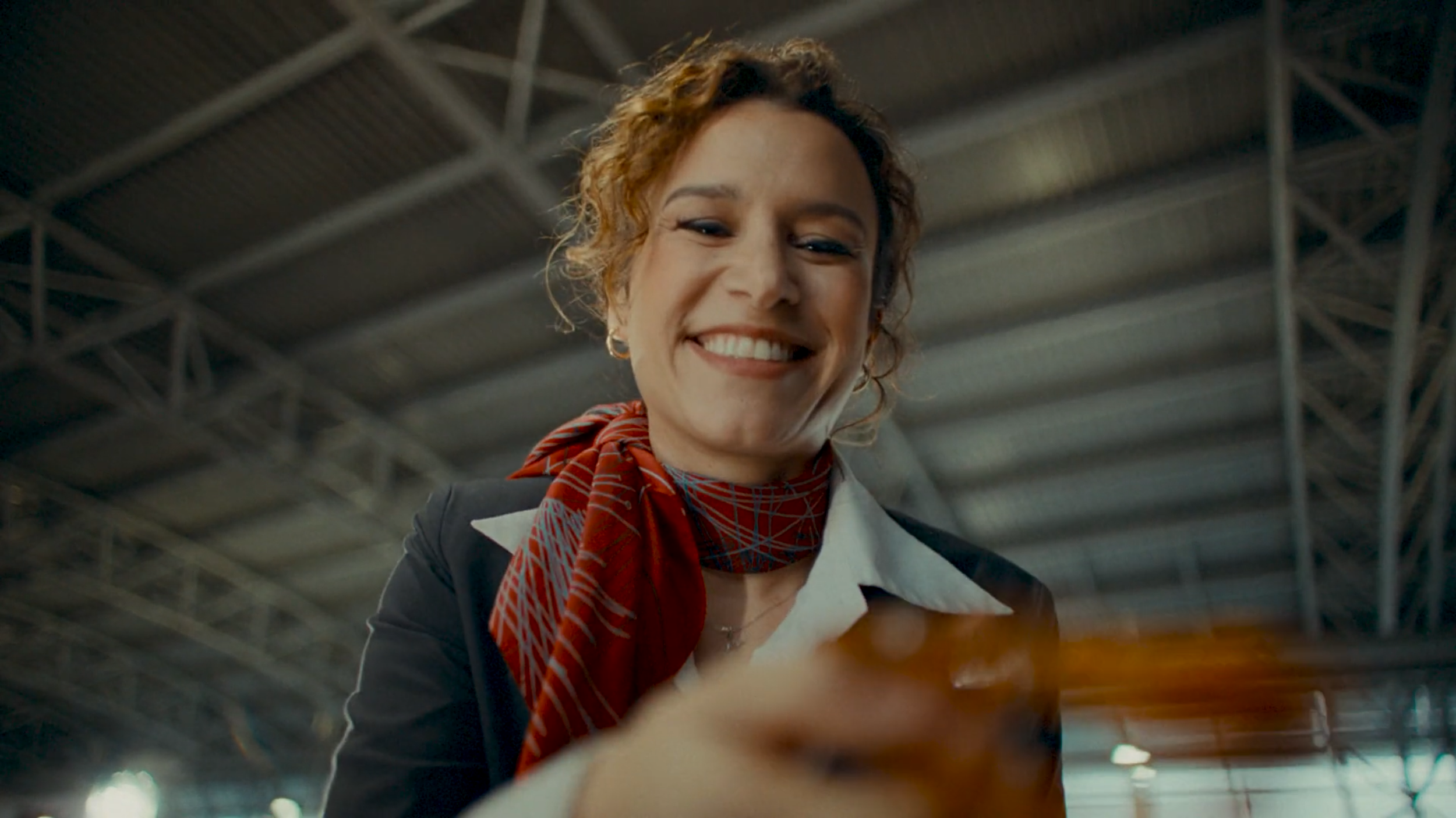 A smiling woman in formal attire, with a red patterned scarf, holding and looking at her phone in an indoor industrial setting.