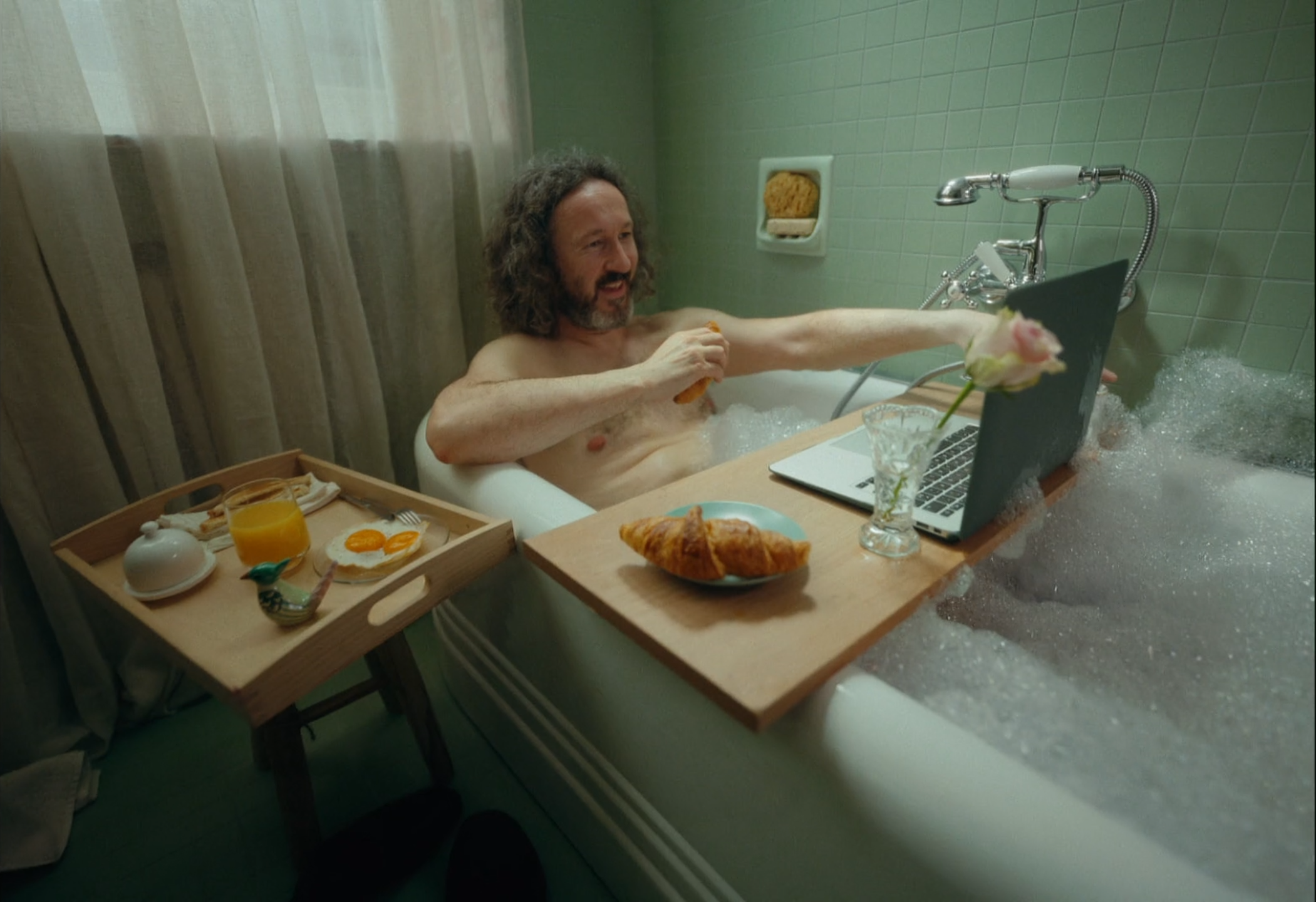 Man with long hair and beard relaxing in a bubble bath with a laptop, breakfast tray, and a vase with a pink flower.