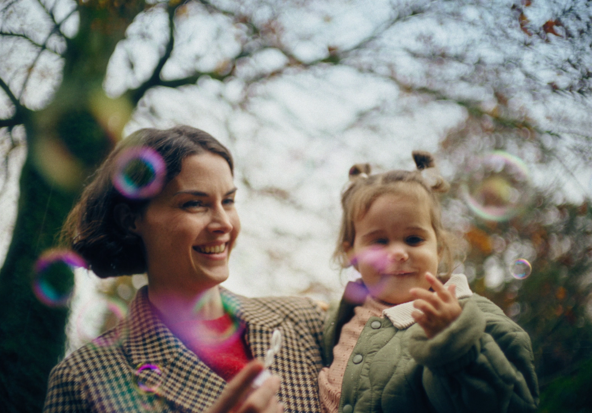 A woman with short dark hair smiling and holding a young girl outdoors, surrounded by colorful soap bubbles, with trees and a cloudy sky in the background.