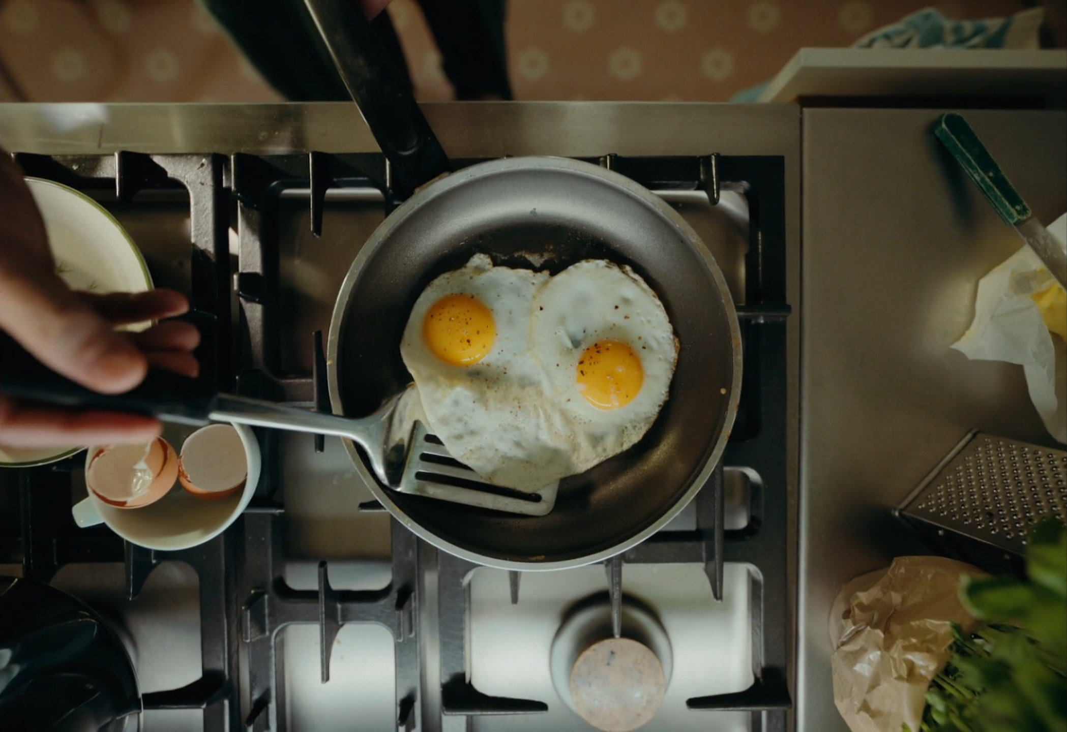 Two eggs frying in a pan on a stovetop, with a hand adding salt.