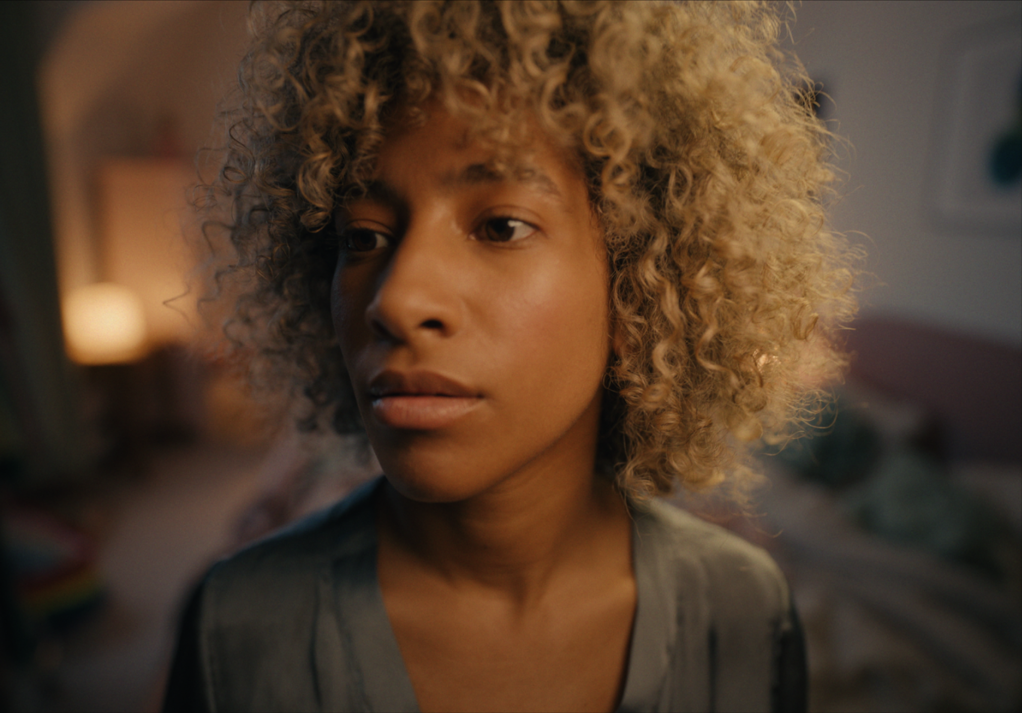 Close-up of a woman with curly blonde hair and tan skin, looking thoughtfully to the side, in a softly lit indoor setting.