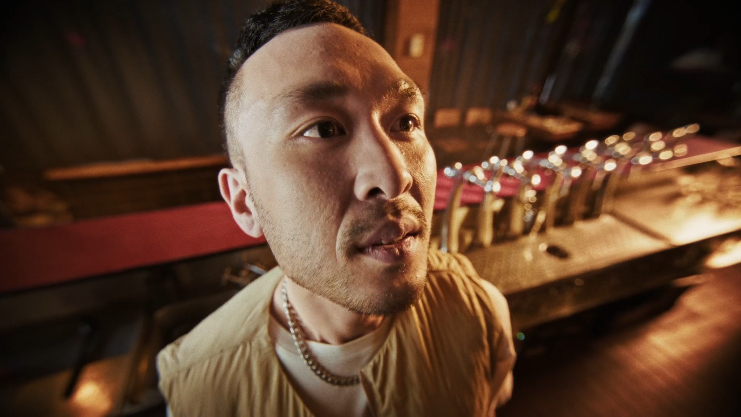 Close-up of a man with short dark hair and a tan shirt in a dimly lit room with shelves and metal chairs in the background.