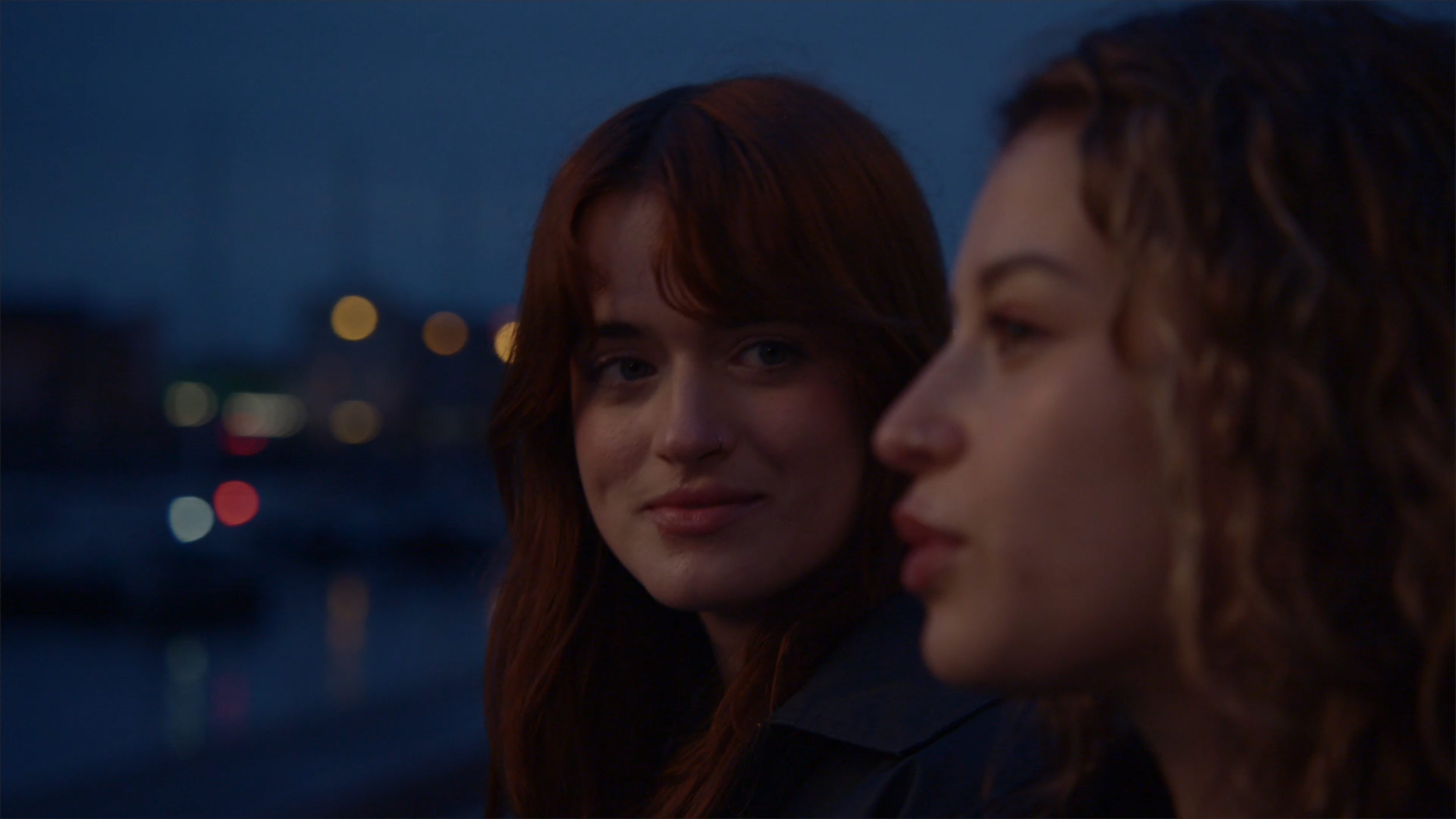 Two women with red hair sitting outdoors in the evening, with city lights blurred in the background.