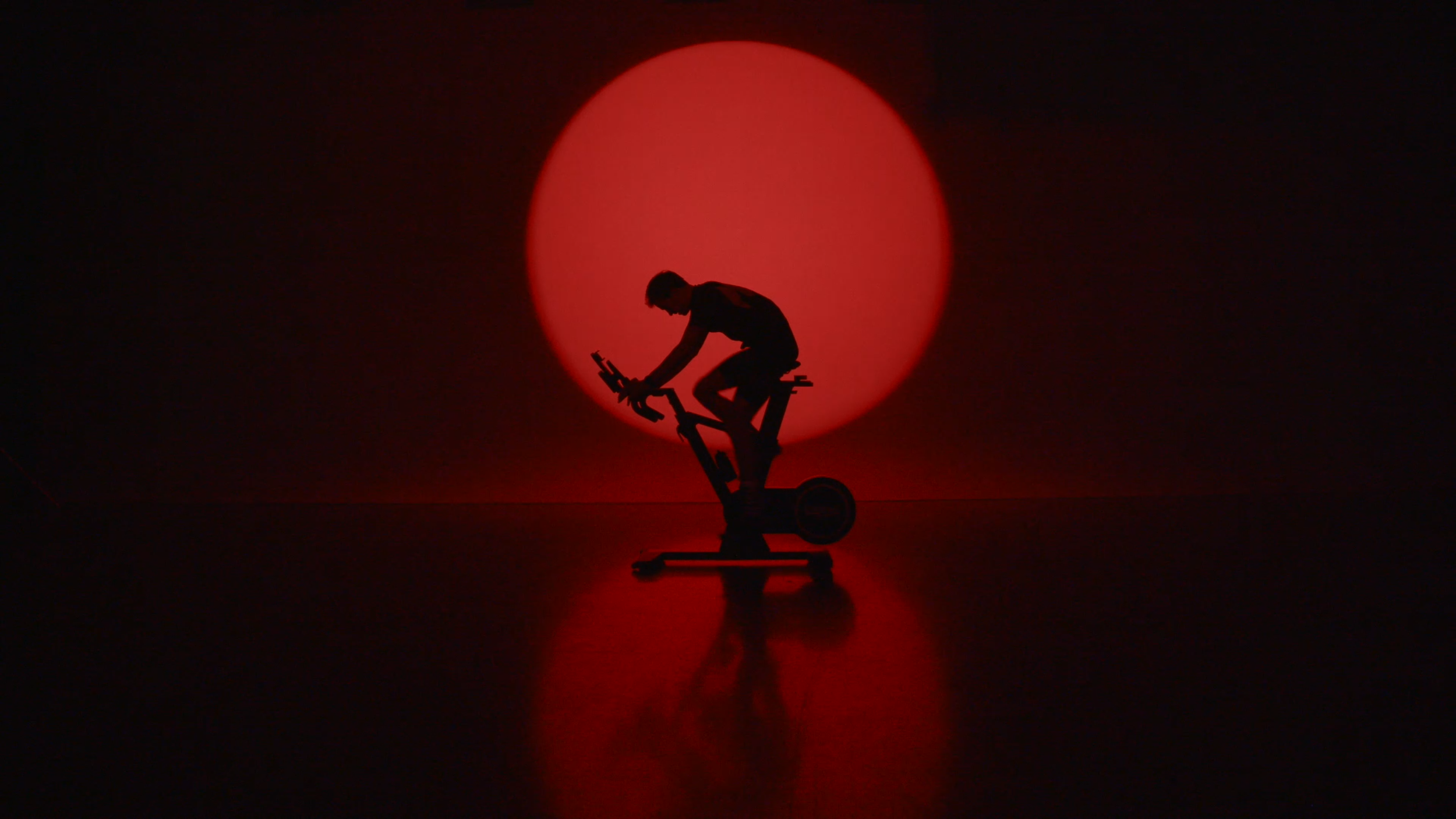 Silhouette of a person riding a stationary exercise bike against a large, bright red circular light in a dark room.