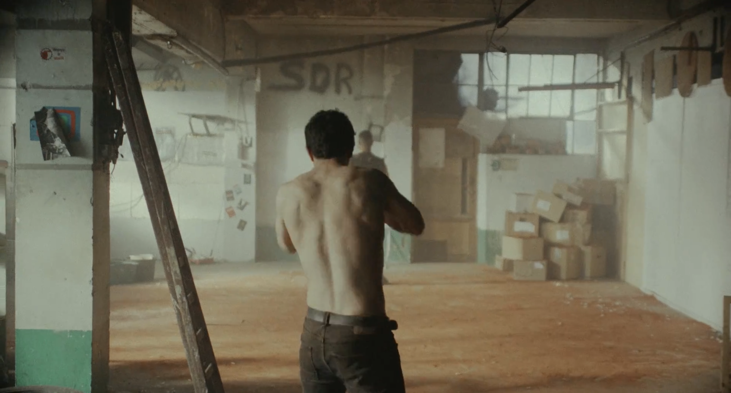 A shirtless man with dark hair stands with his back to the camera in an abandoned or unfinished building filled with dust and debris, with cardboard boxes and construction materials in the background.