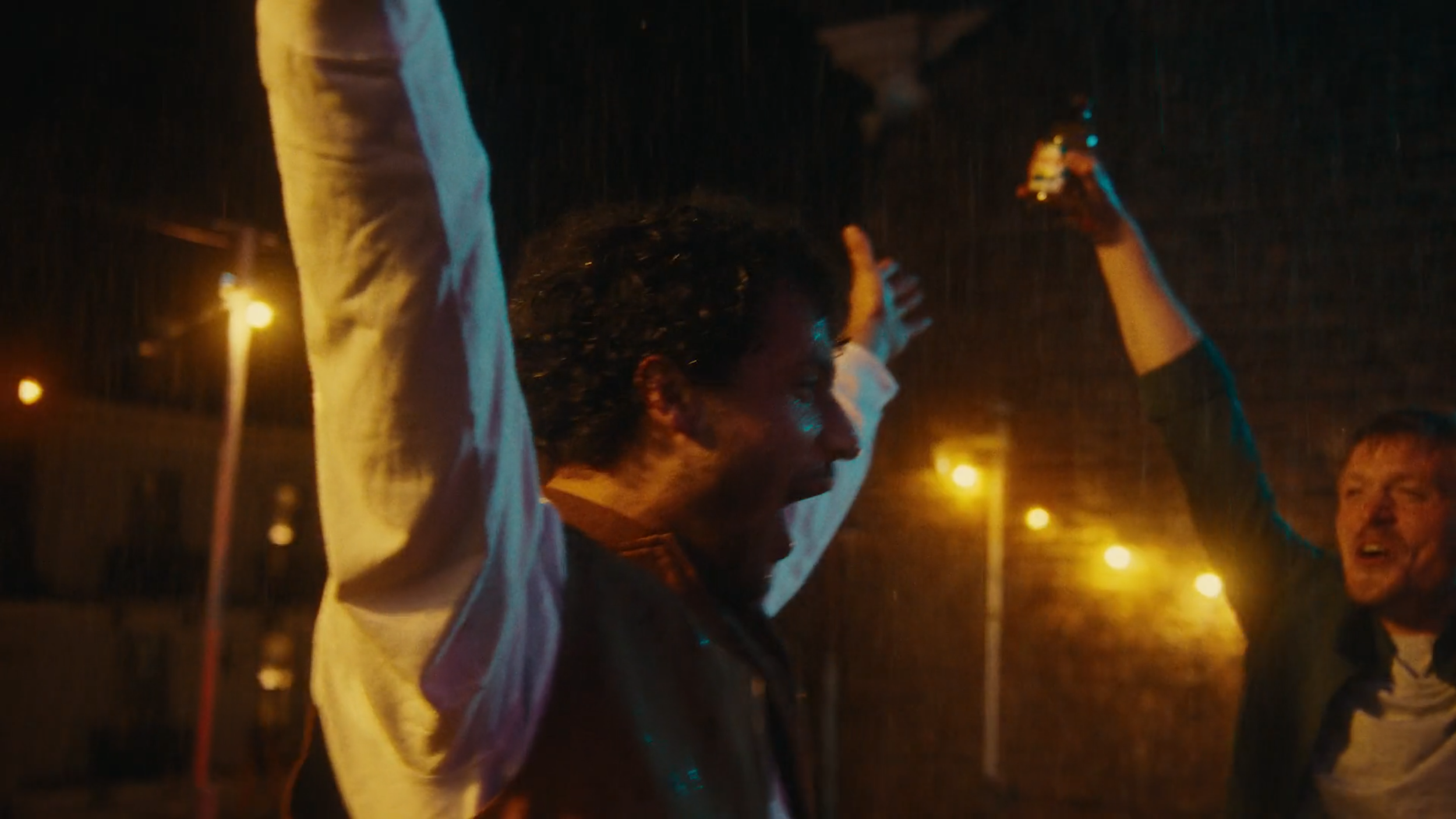 Two people celebrate joyfully in the rain at night, with streetlights illuminating the background.