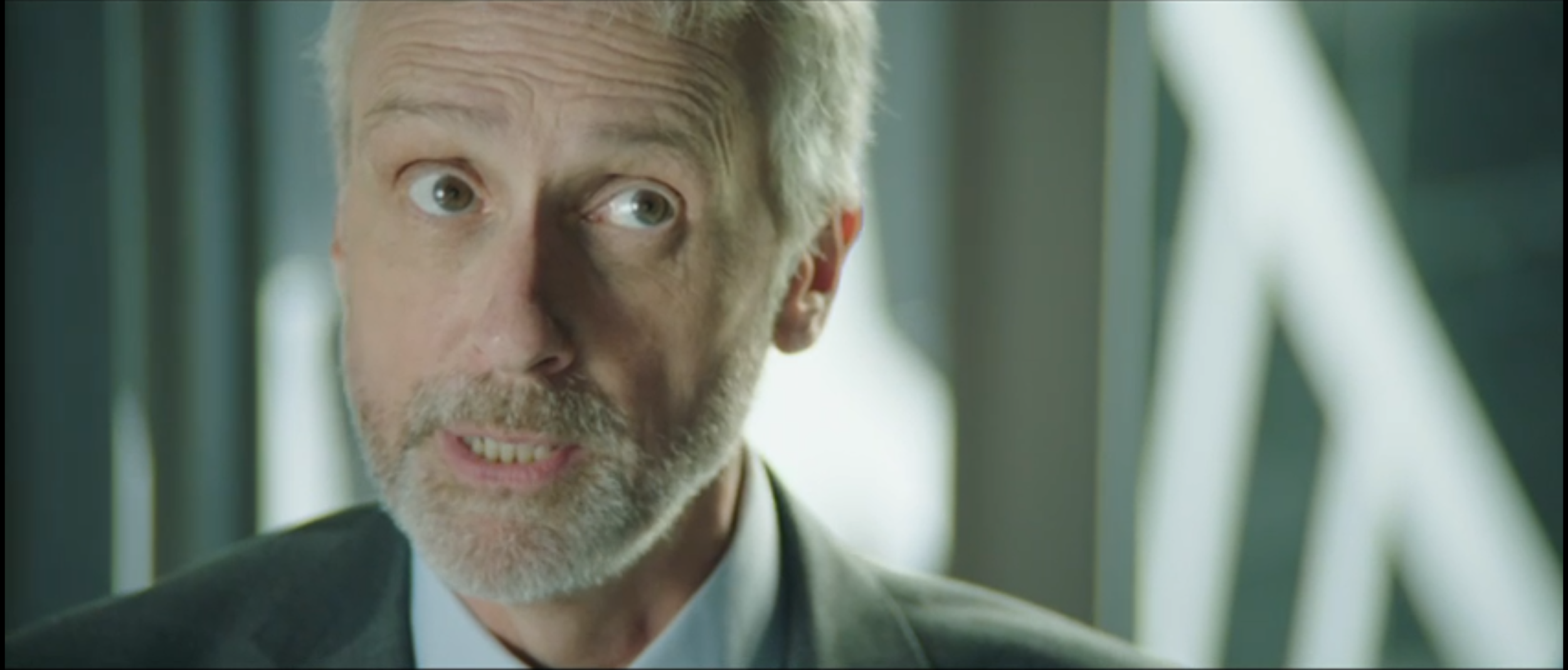 Close-up of a mature man with gray hair, beard, and mustache, wearing a suit, looking to the side indoors with out-of-focus background.