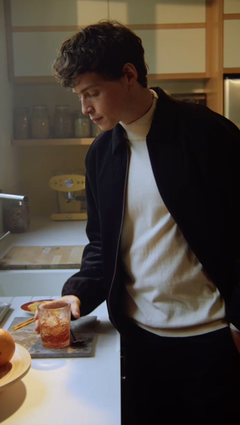 A young man with curly brown hair, wearing a black jacket over a white shirt, is in a kitchen preparing a drink on the countertop. There are jars on the shelves and a yellow coffee machine in the background.