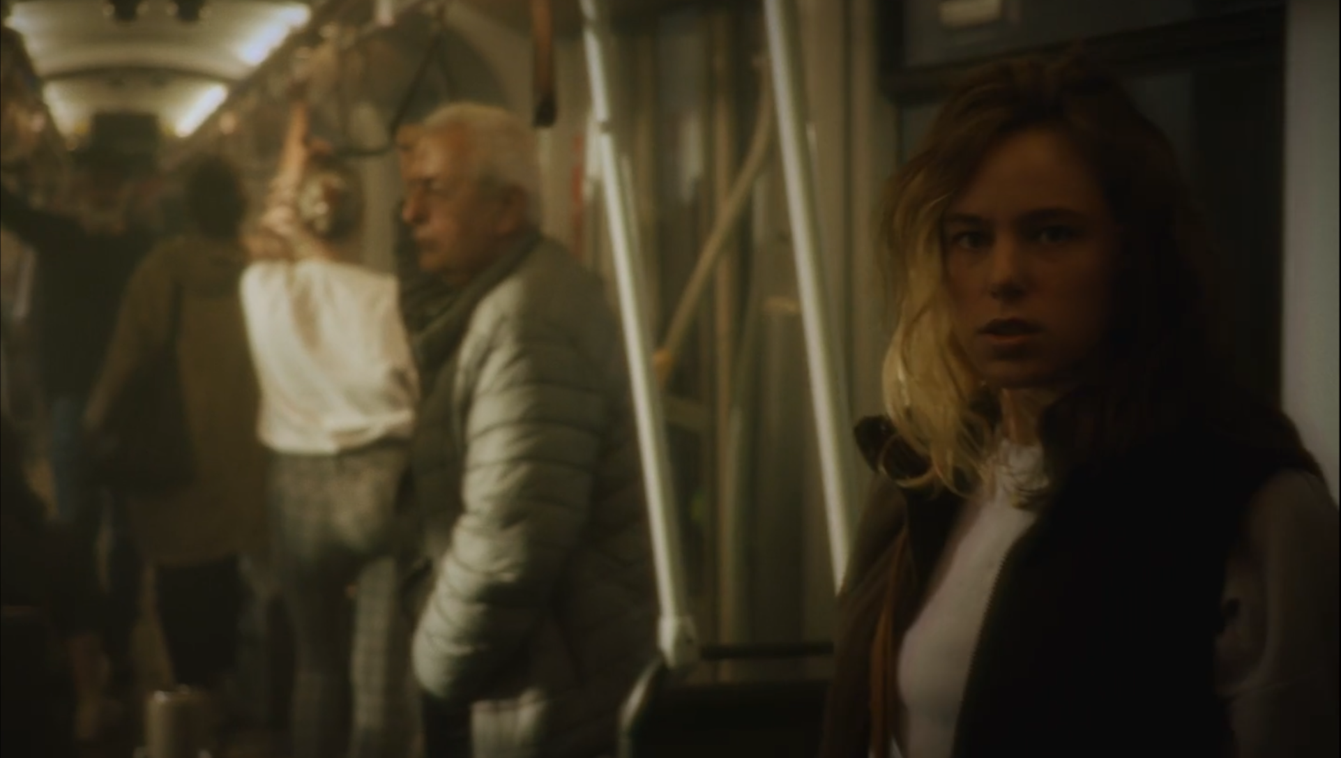 A woman with wavy blonde hair looking to the side inside a dimly lit subway train, with other passengers in the background.