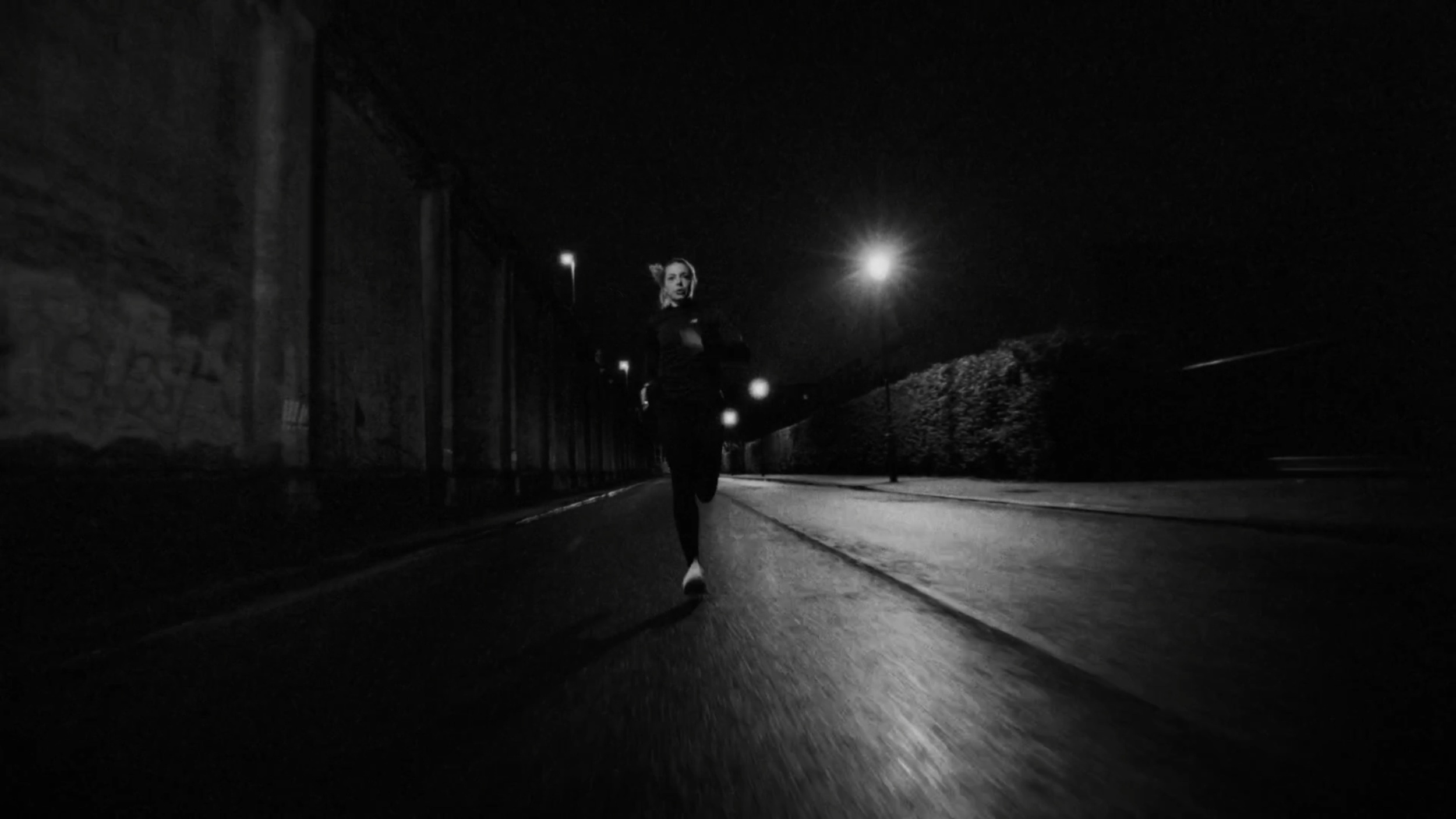 A woman running alone on an empty street at night under streetlights, photographed in black and white.