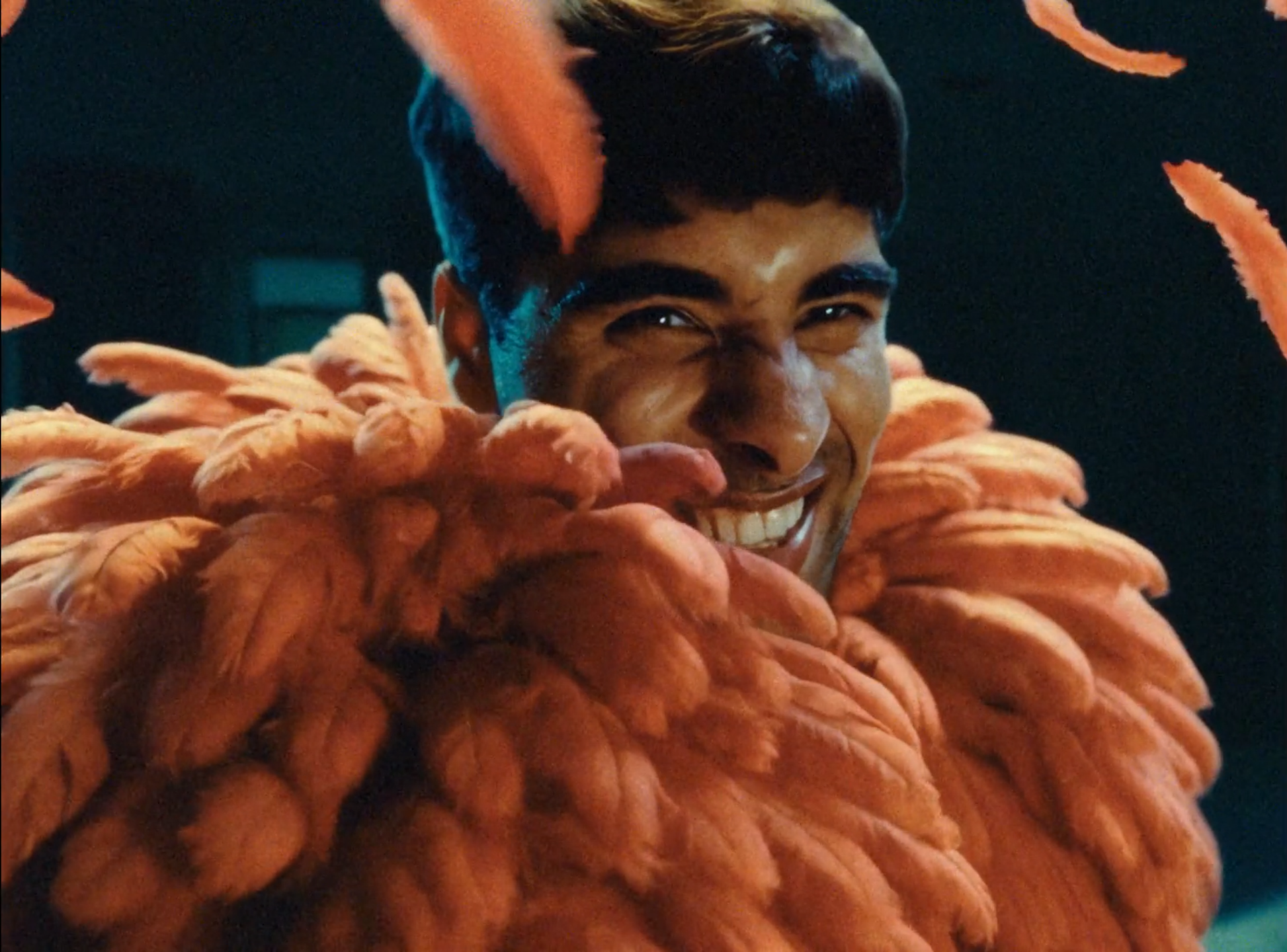 A young man smiling brightly, wearing an orange feathered costume, close-up shot with a dark background.