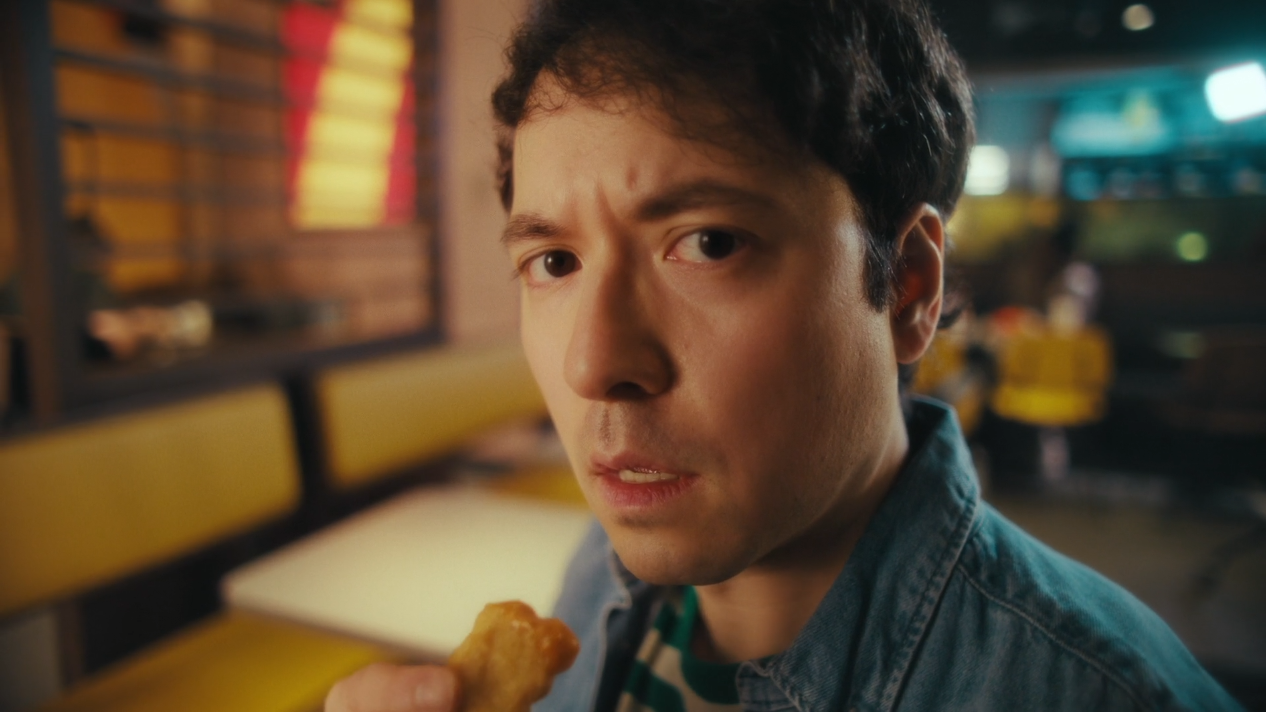 A man with dark hair and a denim jacket looking at the camera while holding a piece of fried chicken inside a fast food restaurant with yellow seating and a blurred background.