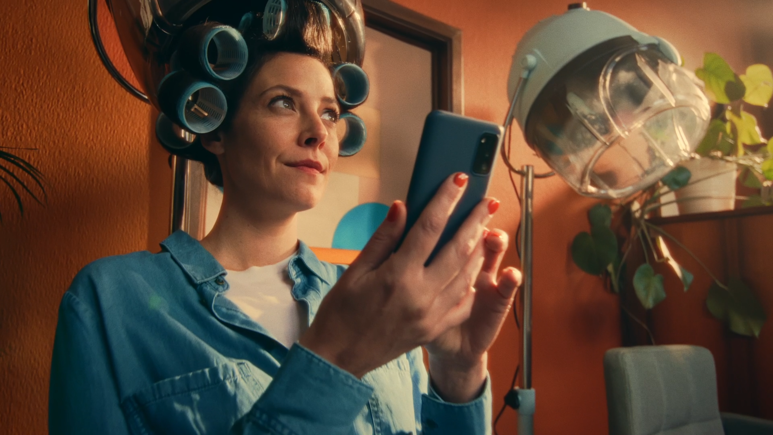 A woman with hair rollers is looking at her smartphone in a room with warm orange walls, a framed picture, and a green plant in a clear pot.