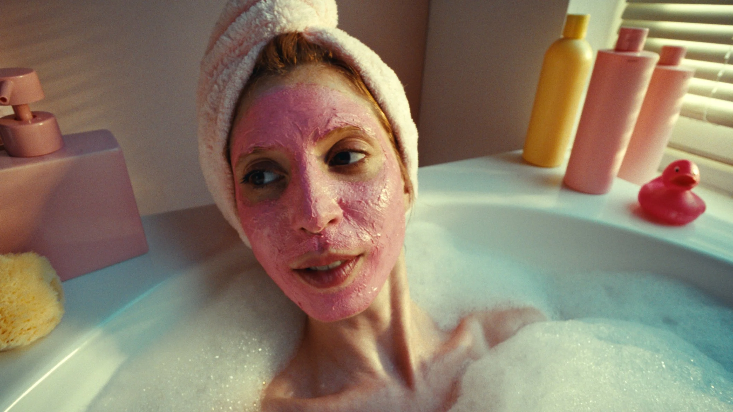 A woman with a towel wrapped around her head is in a bubble bath, covered in bubbles, with pink facial mask on her face, looking relaxed.