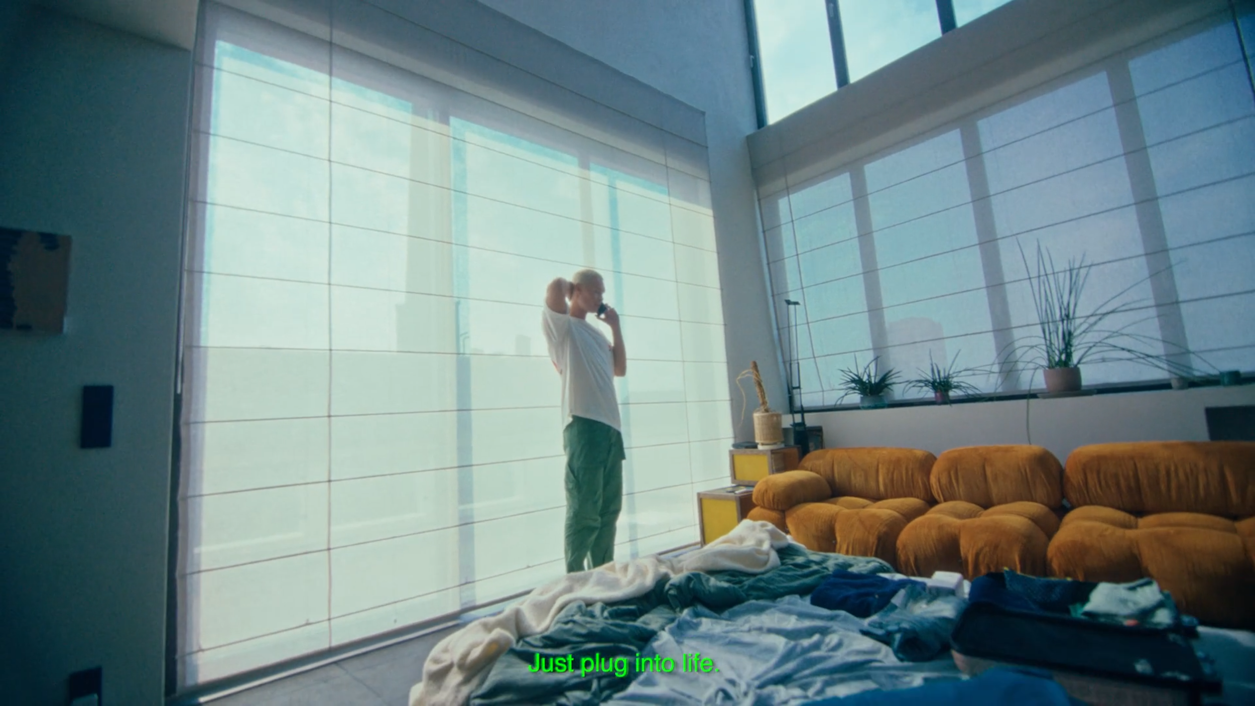 A woman standing near large window blinds in a modern living room, talking on the phone. The room has a mustard-colored sectional sofa, a small yellow side table with a vintage television, and several potted plants on the windowsill. Clothes are scat