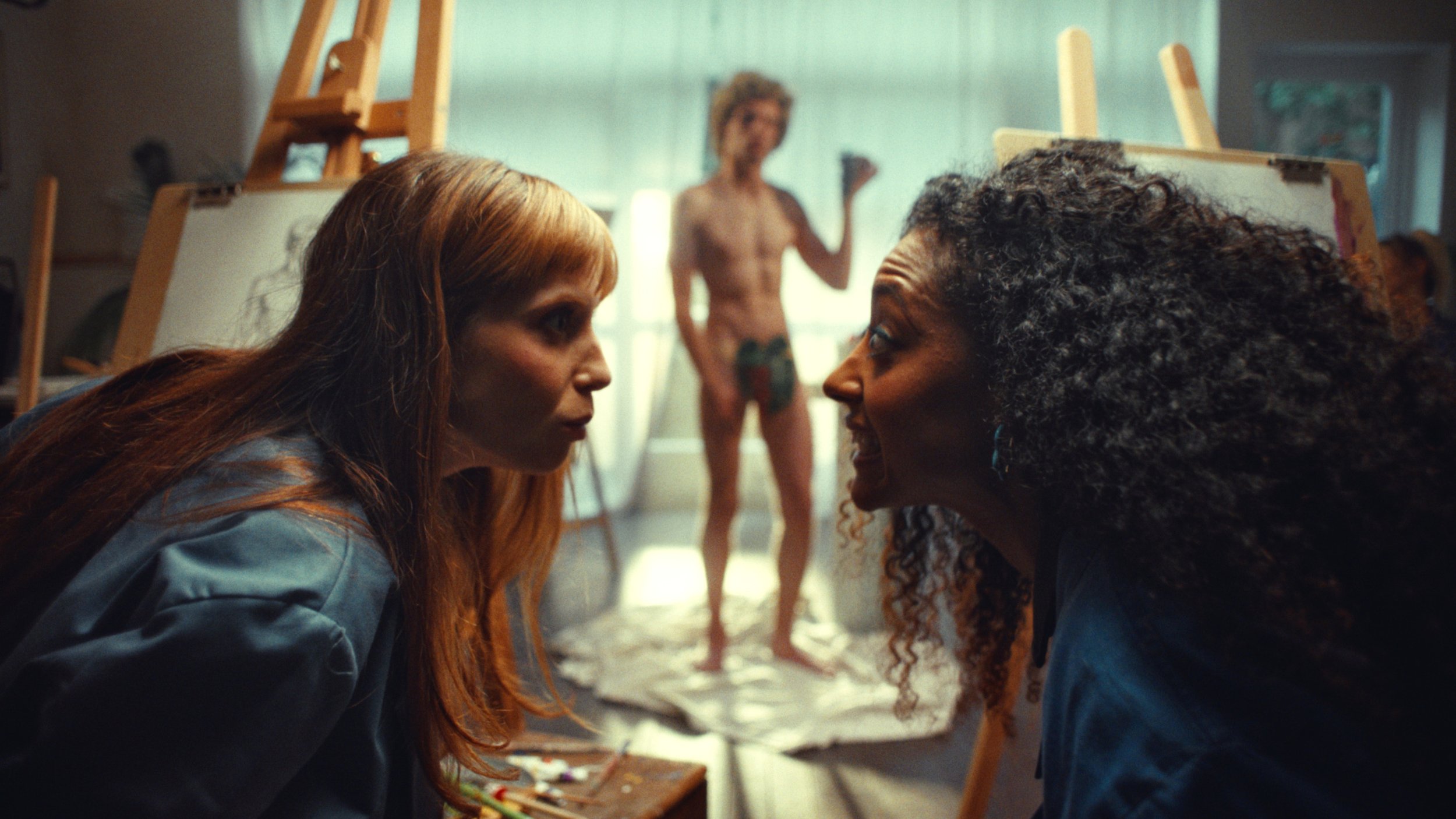 Two women with serious expressions in an art studio, leaning toward each other. In the background, a shirtless man stands on a canvas with paint smudges, holding a paintbrush, while art supplies and easels are visible.