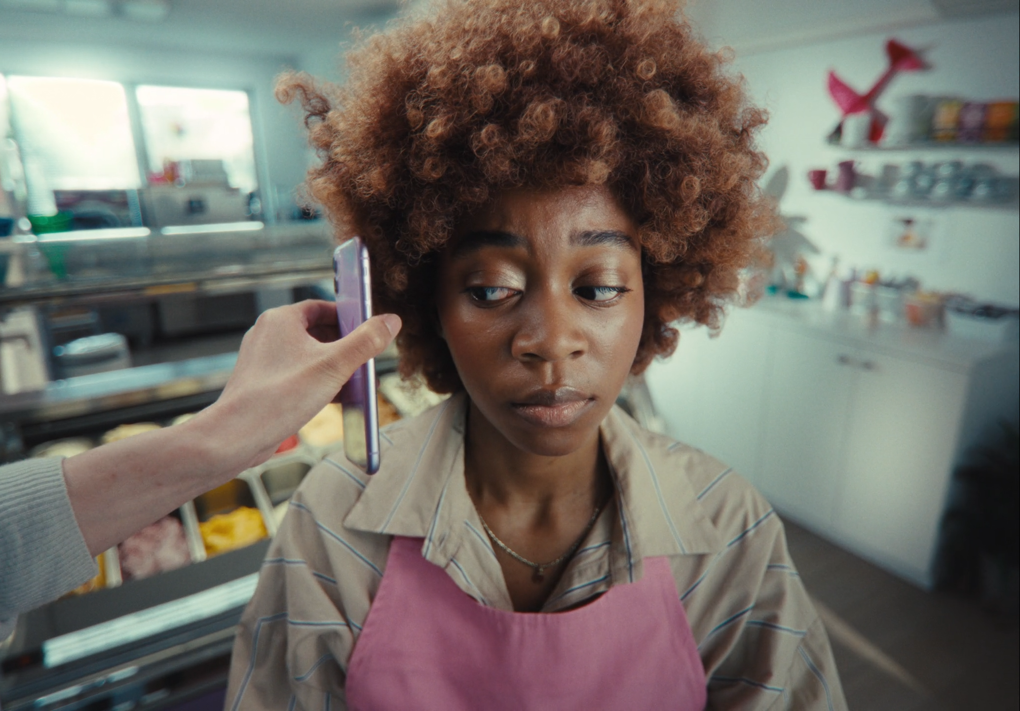 A young woman with curly hair wearing a beige striped shirt and pink apron sitting in a kitchen, while someone holds a smartphone to her ear.