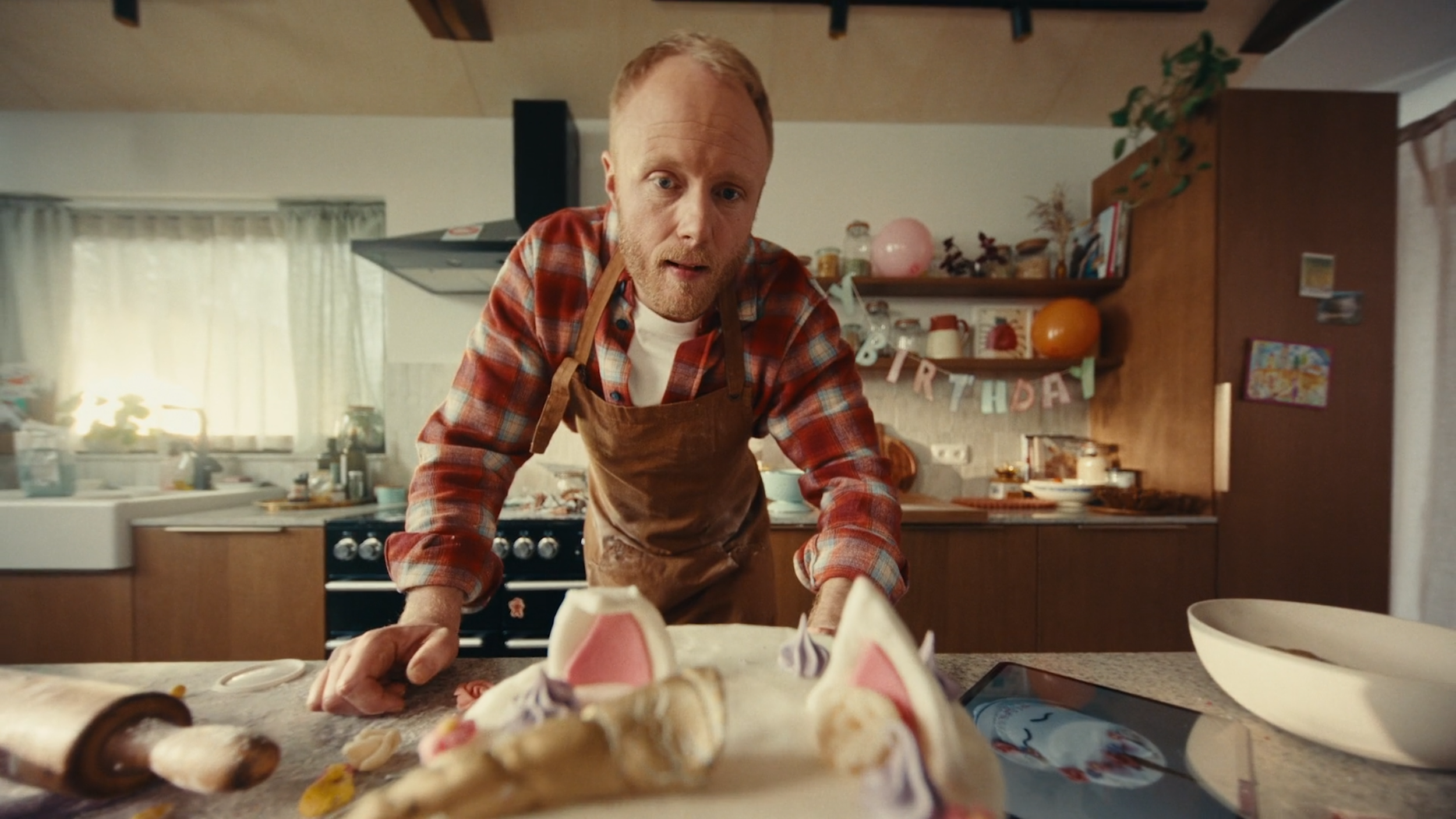 A man with light hair and a beard leaning over a table with a partly eaten birthday cake with pink bunny ears, in a kitchen decorated for a birthday celebration.