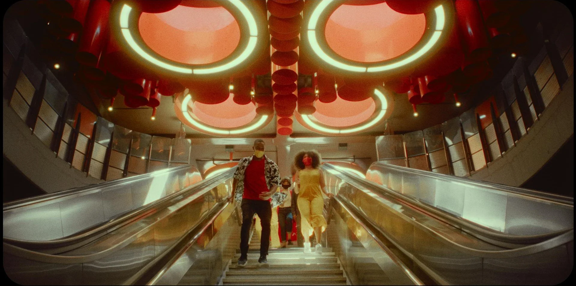 People riding an escalator in a modern, illuminated indoor space with bold red and neon ceiling decorations.