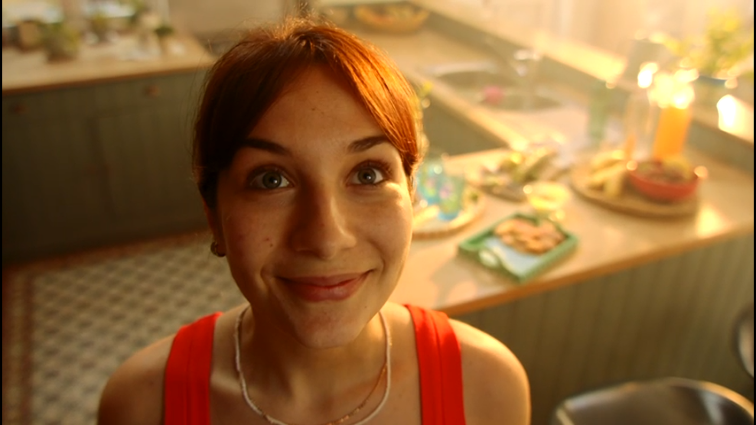 A woman with short reddish-brown hair smiling in a kitchen with a blurred countertop and various food items in the background, illuminated by warm sunlight.