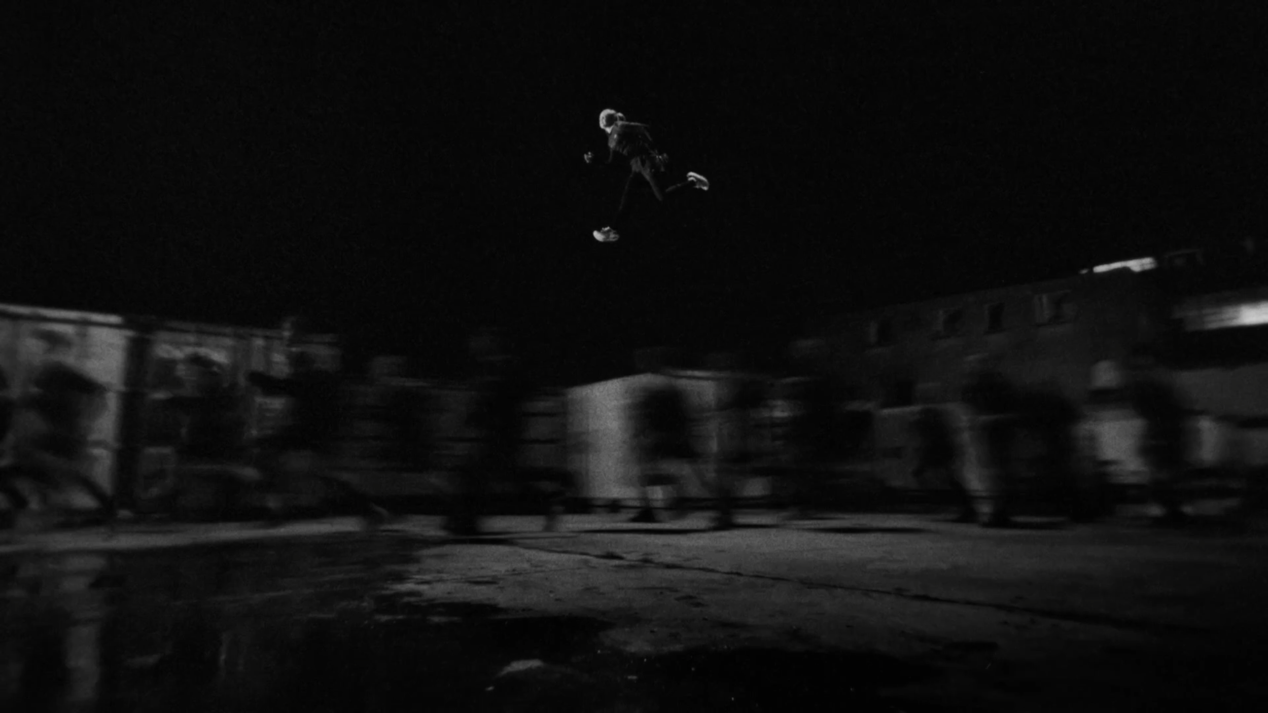 A person performing a jump on a skateboard at night, with blurred figures and buildings in the background.