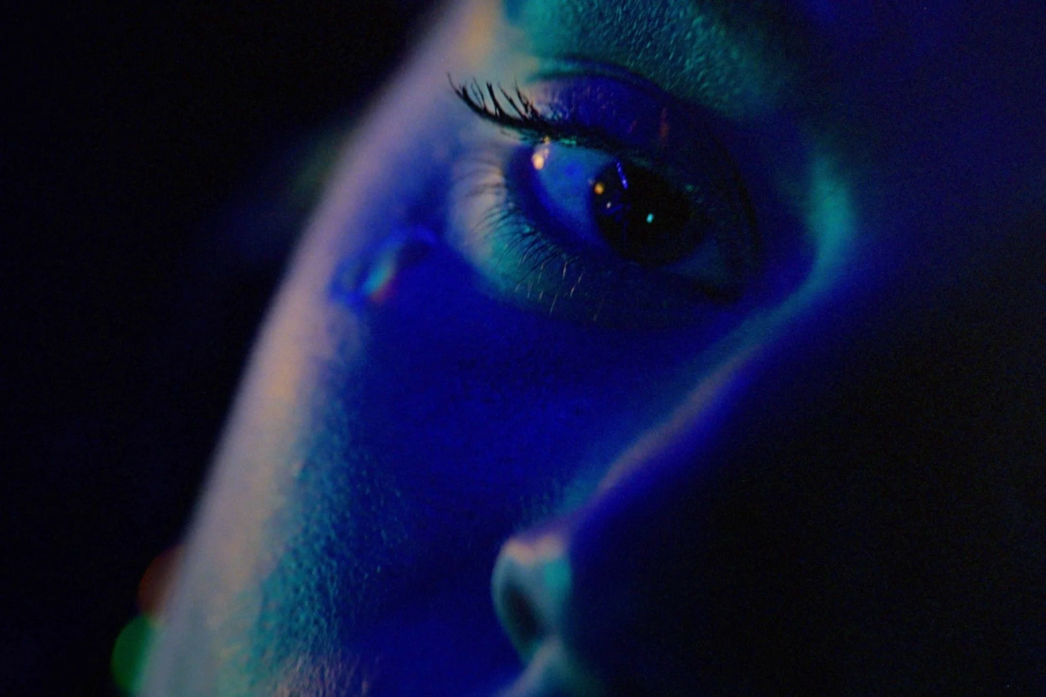 Close-up of a woman's eye illuminated by blue and green lights, with long eyelashes and reflections in the iris.