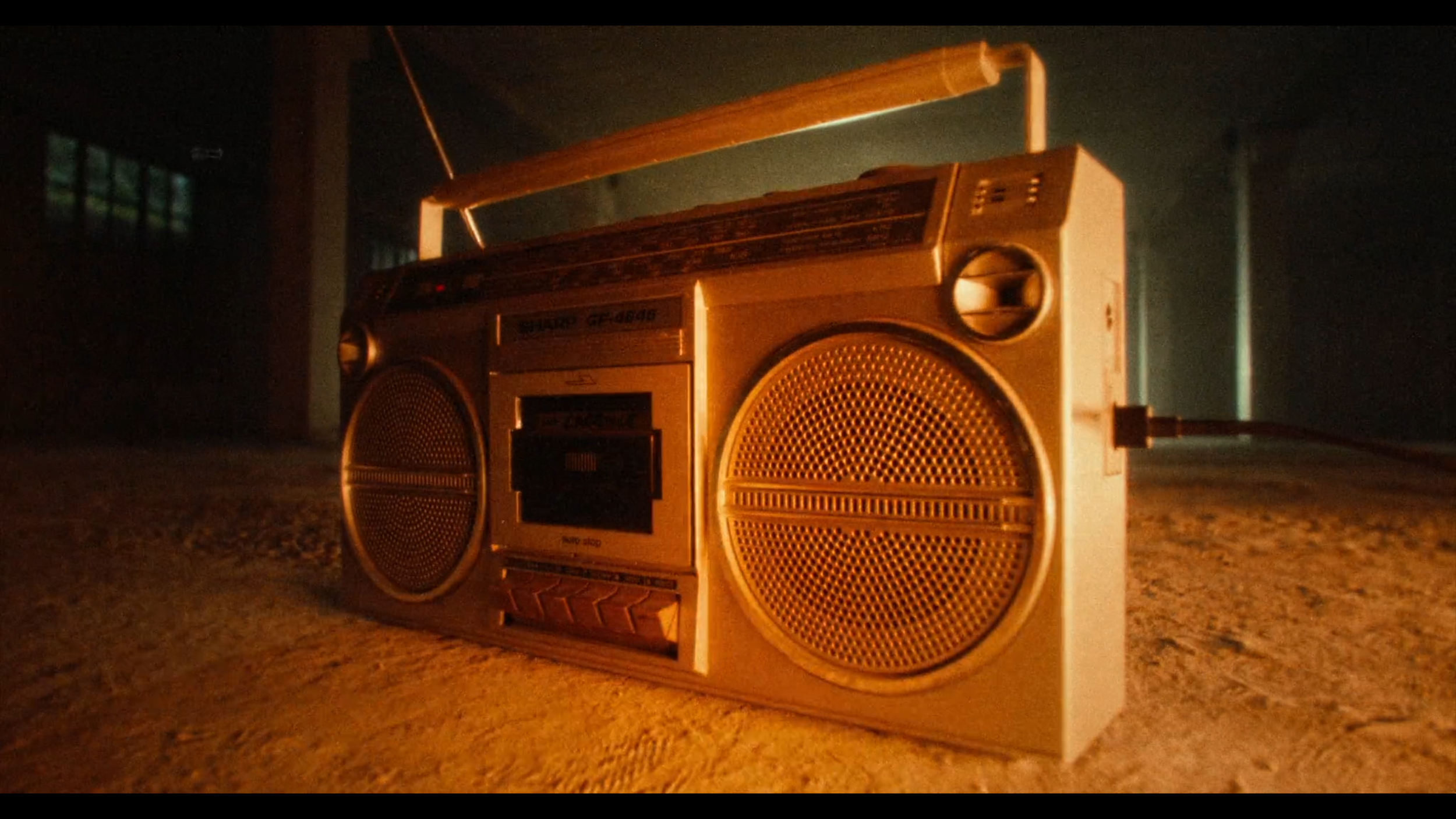An old silver boombox with two speakers on the front, a cassette tape deck, and an antenna, placed on a textured surface in a dimly lit room.