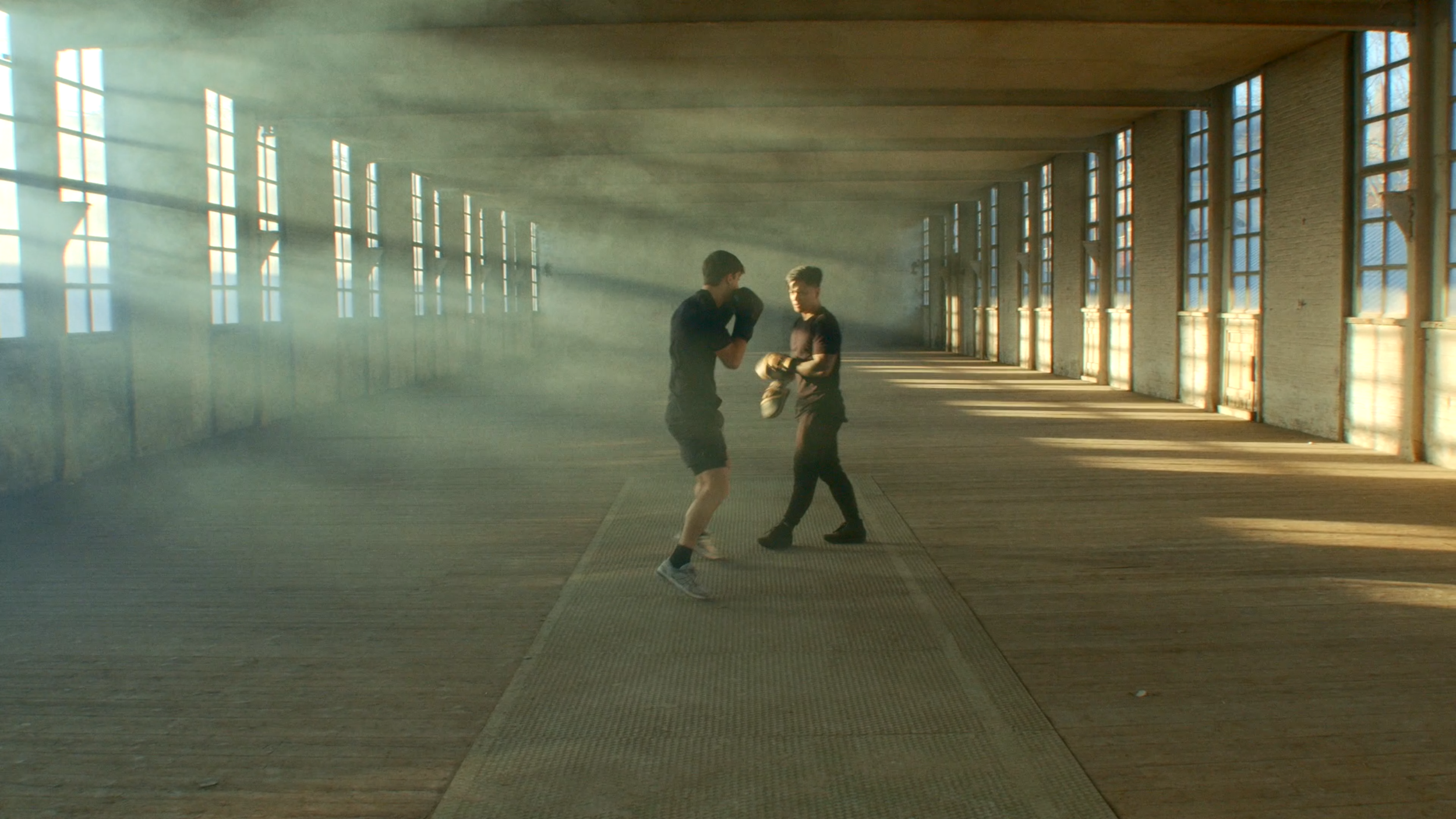 Two young men practicing boxing in a spacious, sunlight-filled gym with large windows and wooden floors.