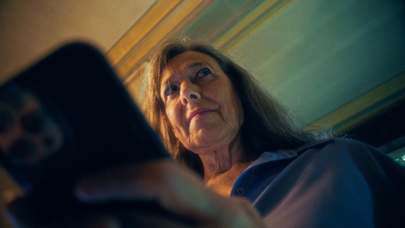 An elderly woman with long gray hair looking down at her phone with a stern expression in a room with wooden crown molding.