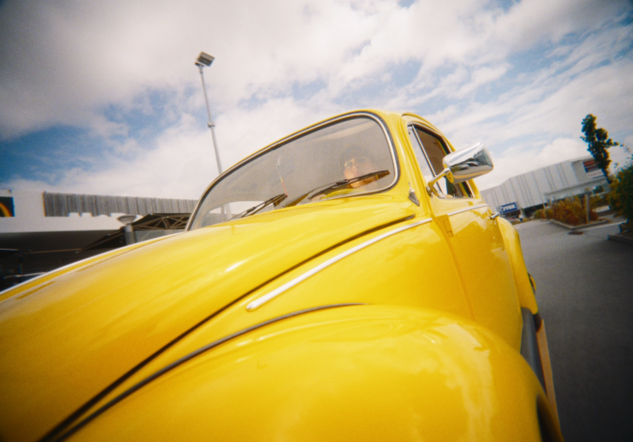 Close-up of a yellow vintage car, taken from the front, with a person inside wearing sunglasses, against a partly cloudy sky with a parking lot and buildings in the background.