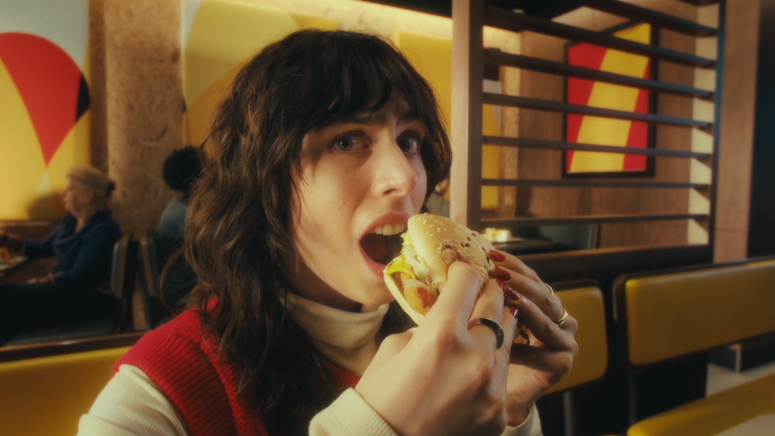 A woman with dark wavy hair, wearing a red and cream-colored sweater, is sitting in a restaurant and eating a burger.