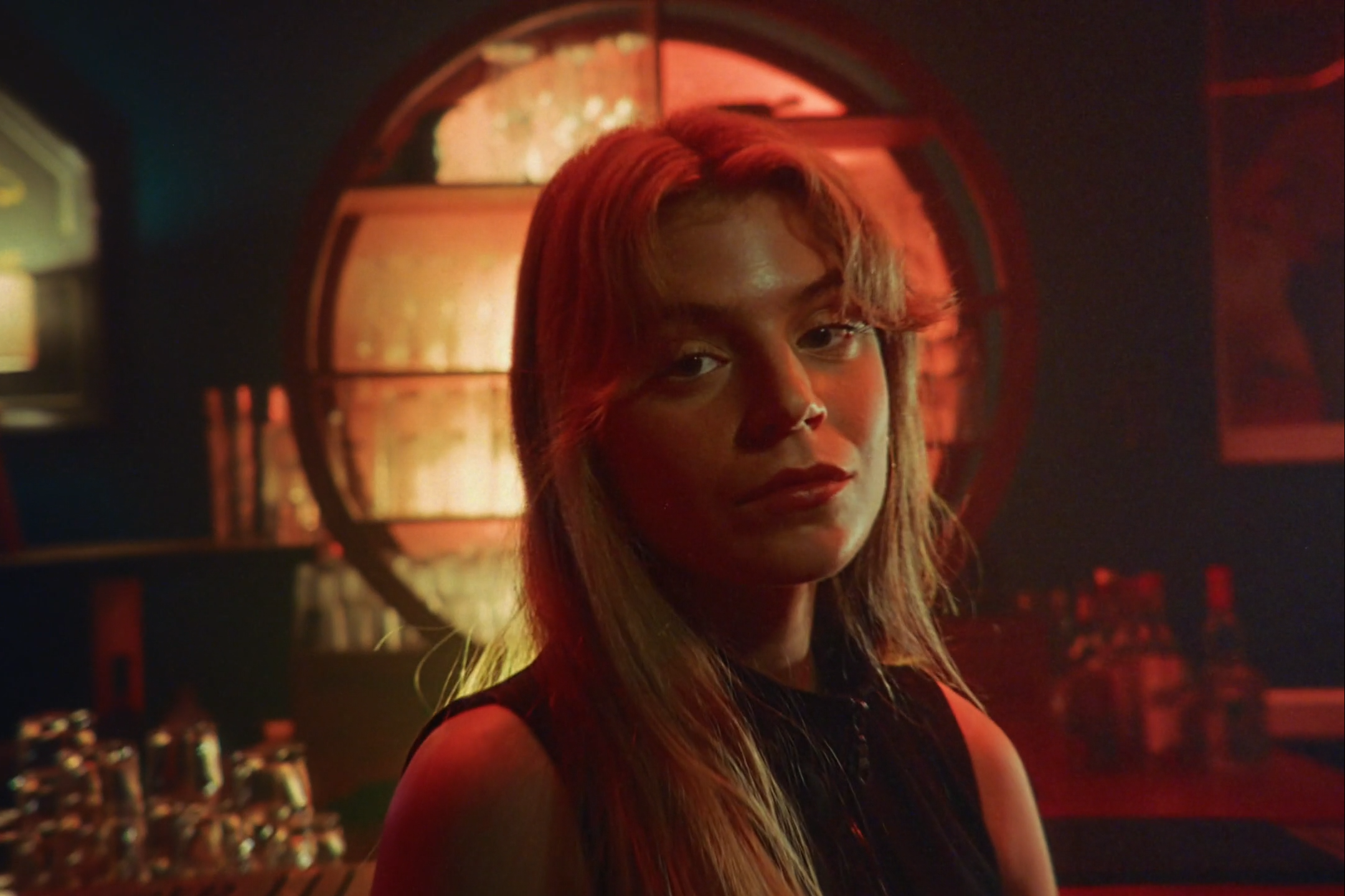 A young woman with light hair and a dark top in a dimly lit room with a red glow, looking at the camera with a slight smile, behind her a stained-glass window and bottles on a bar.