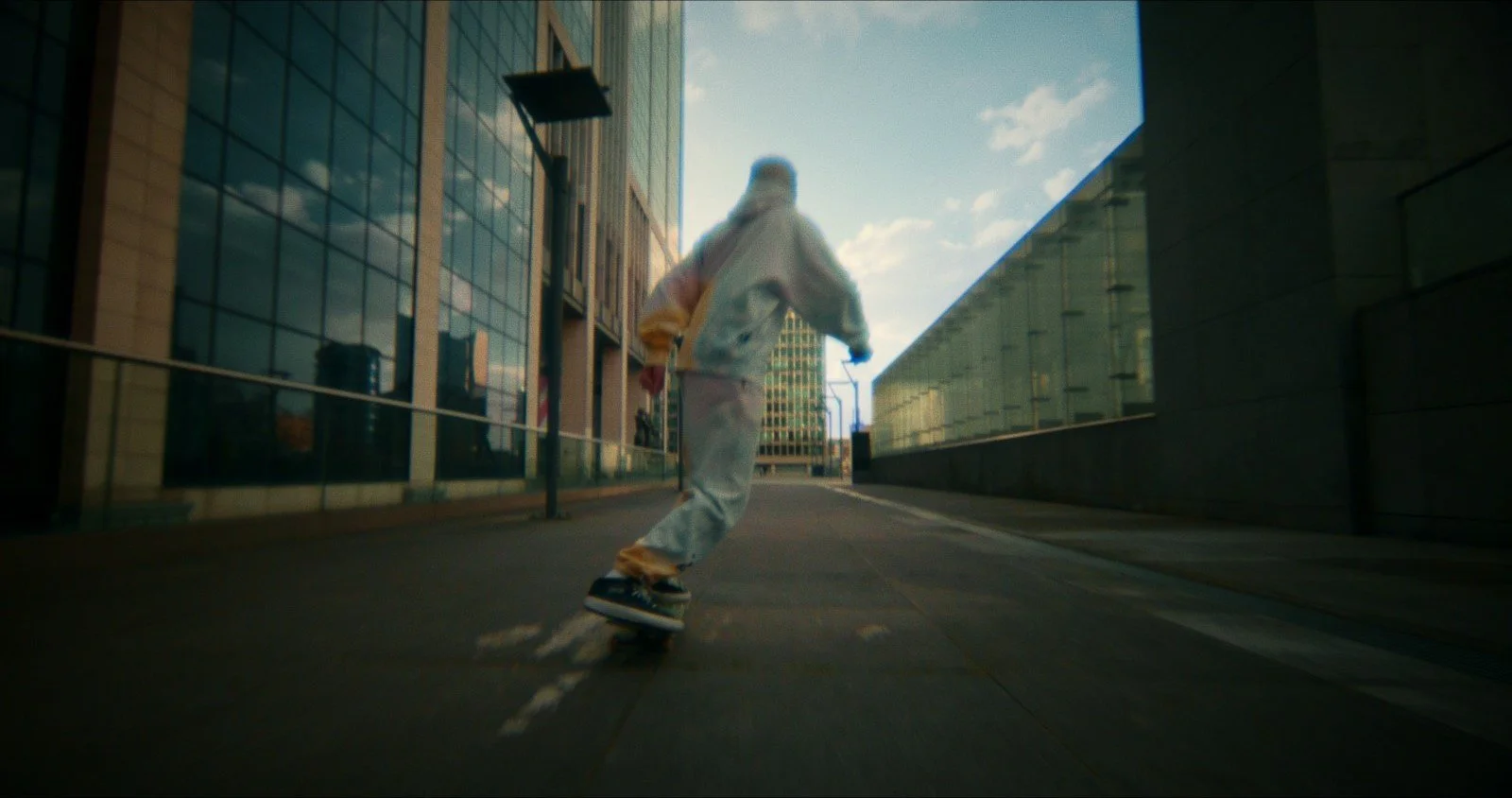 Person skateboarding down an urban street during daytime, surrounded by modern glass buildings.