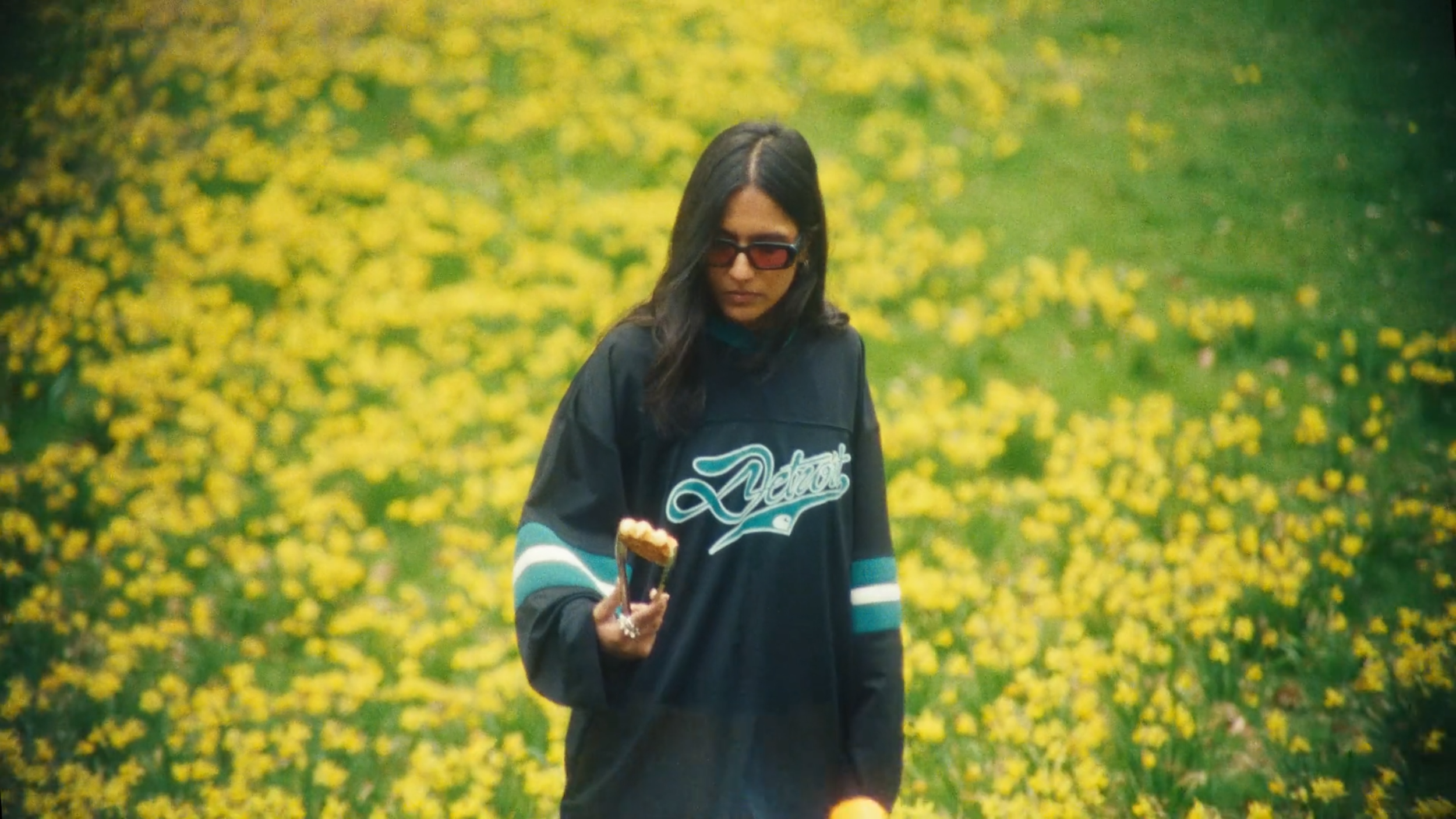 A woman with long dark hair wearing glasses and a dark sports jersey with light blue and white stripes on the sleeves, standing in a field of yellow flowers, holding a piece of cake in her right hand.