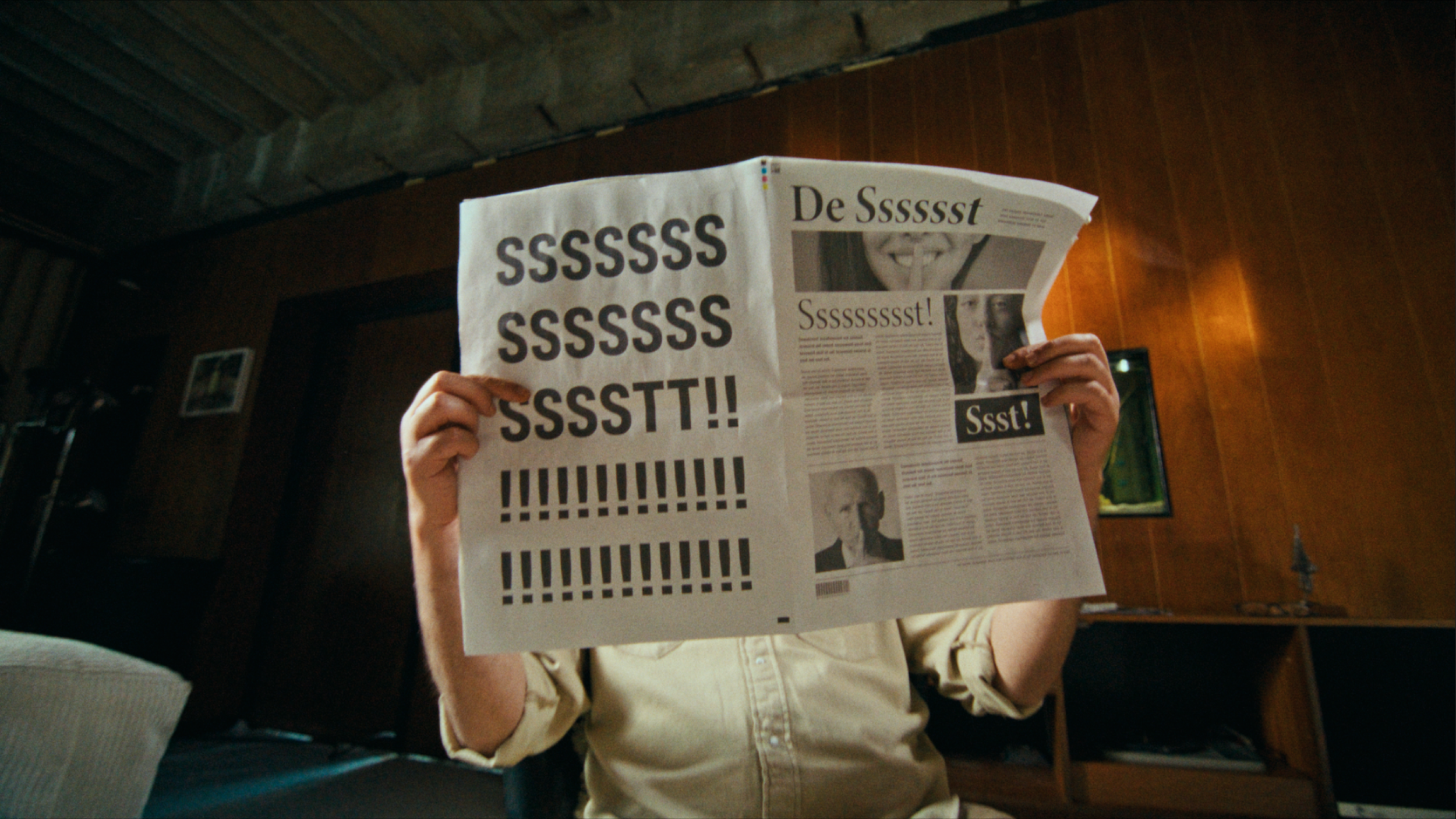 A person holding a newspaper with large black 'S' characters and exclamation points, obscuring their face, in a room with wooden walls and a couch.