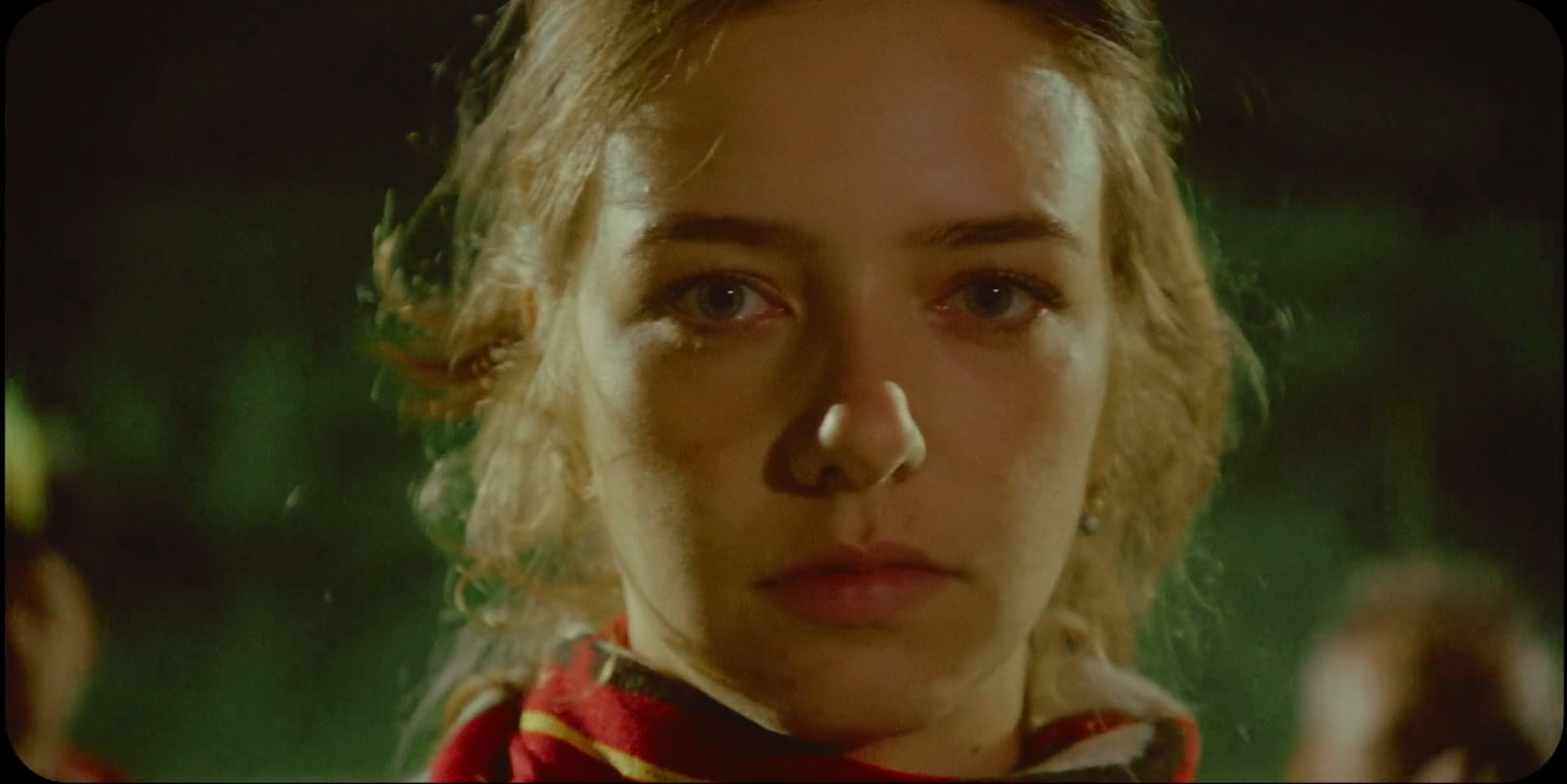 Close-up of a young woman with short, wavy blond hair, looking intently at the camera with a serious expression, in a dimly lit outdoor setting at night.