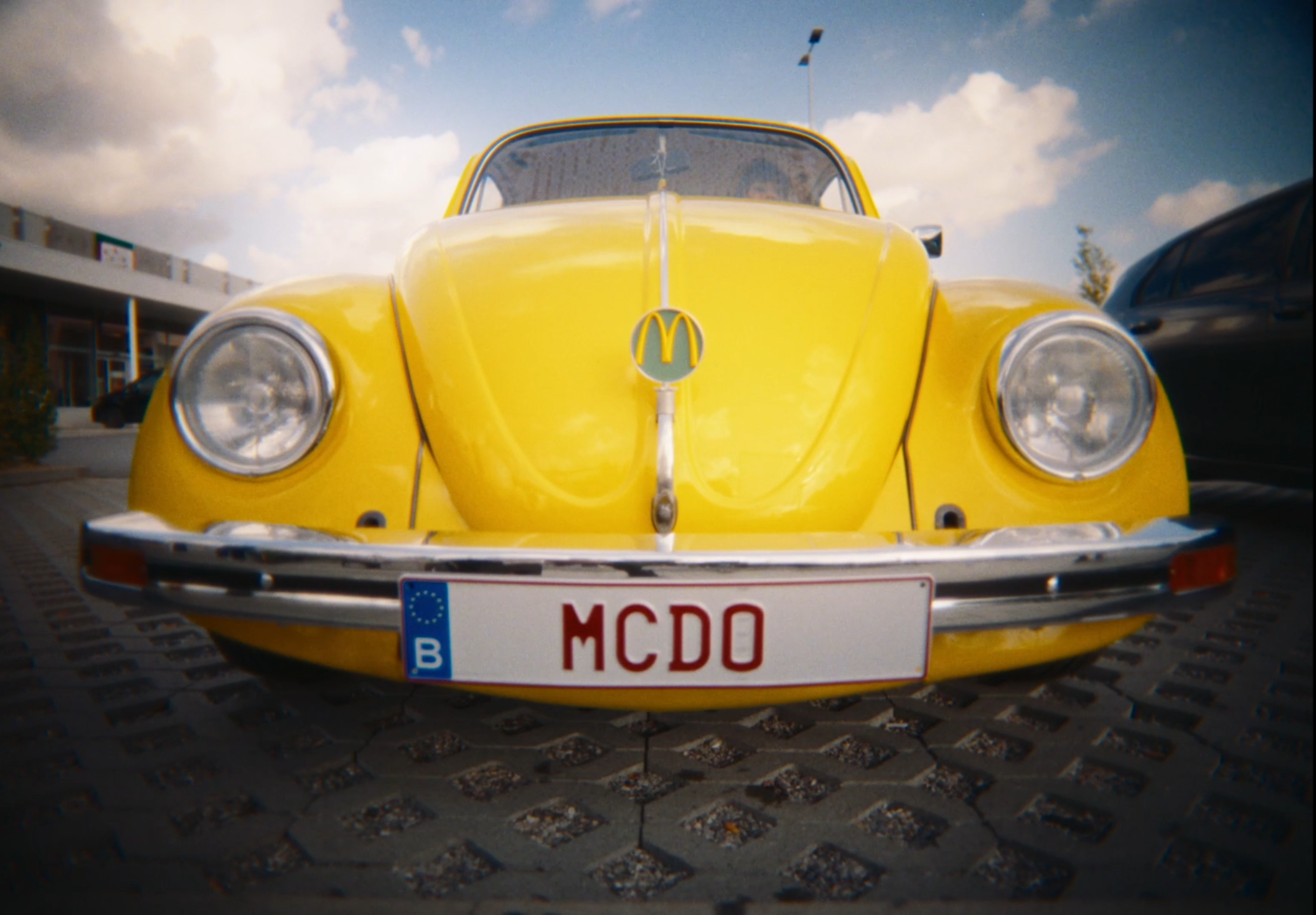 A yellow vintage Volkswagen Beetle with a McDonald's logo on the hood, parked in a lot with a European license plate that reads 'MCDO', under a partly cloudy sky.