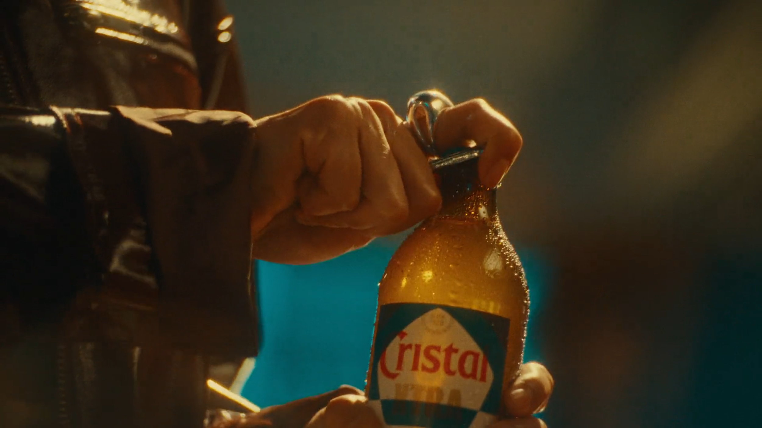 Close-up of a person's hand holding a beer bottle with condensation, wearing a dark leather jacket, in a dimly lit setting.