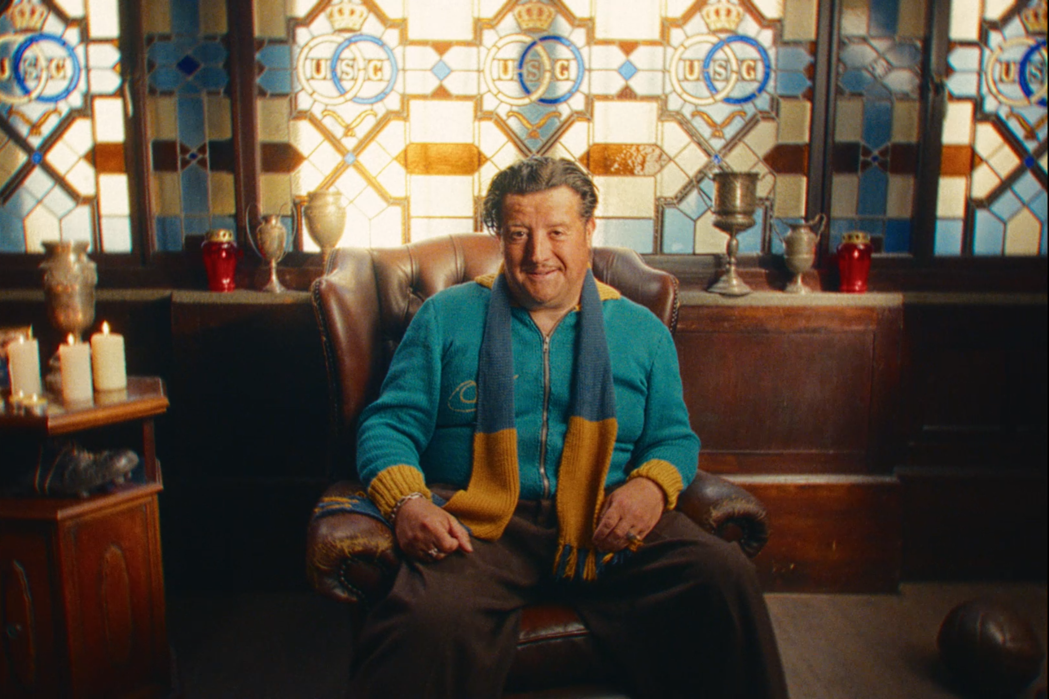 A man with gray hair and a beard sitting in a leather armchair in a room with stained glass windows. He is wearing a blue hoodie with yellow accents and a blue and yellow scarf. The room has candles, vases, and other decorative objects.