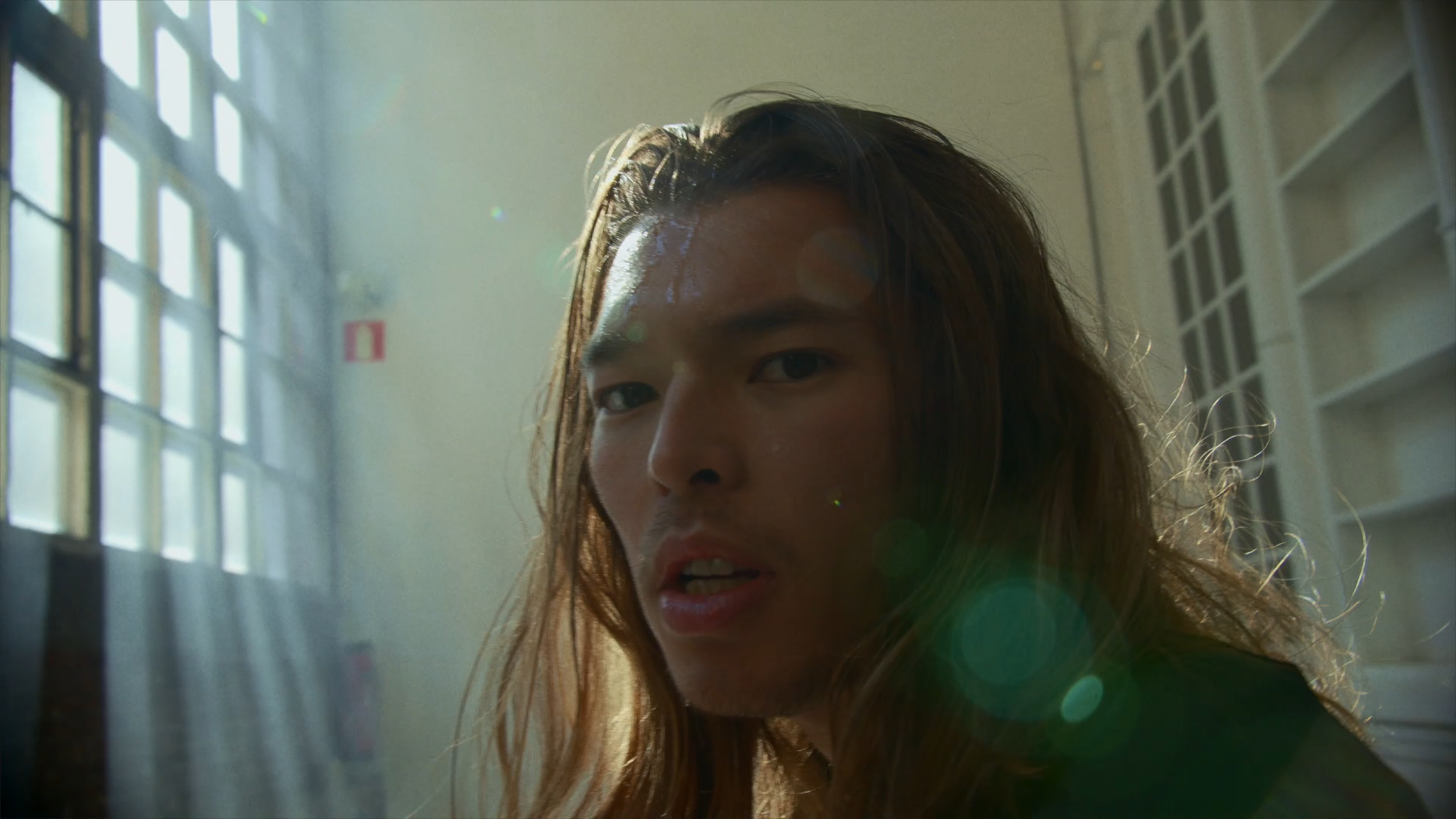 Close-up of a young person with long, wet hair illuminated by sunlight, standing indoors near a window with glass blocks and a bookshelf in the background.