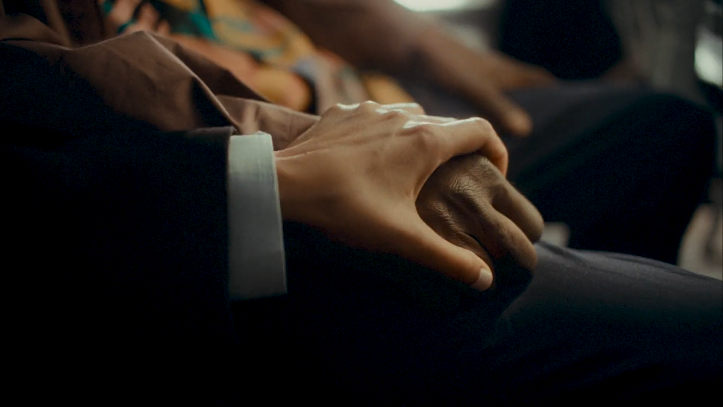 Close-up of two people's hands resting on their laps, one with darker skin and the other with lighter skin, in a comforting or supportive gesture.